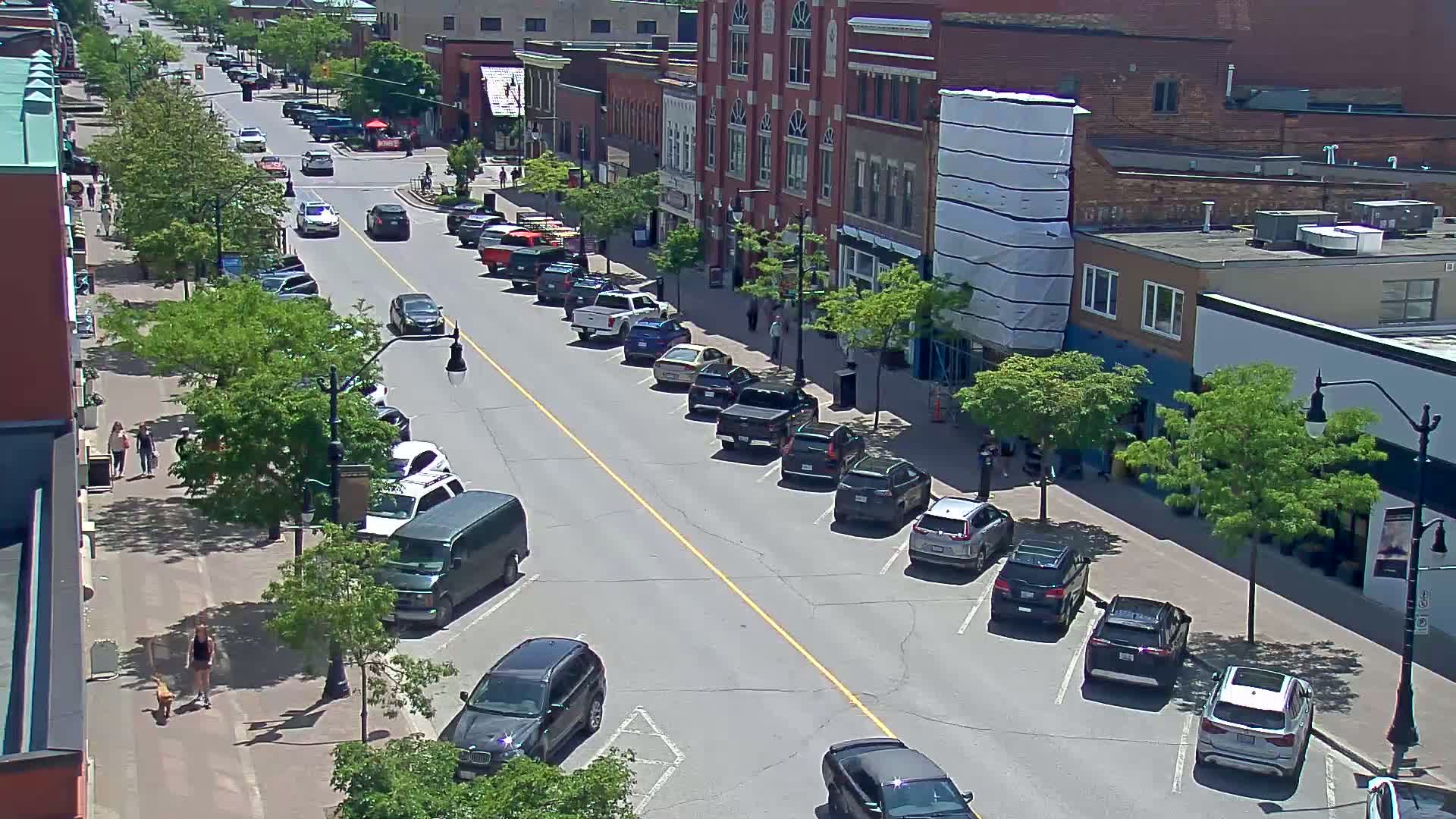 A sunny street scene shows cars parked along both sides of a road in front of brick buildings, with pedestrians walking on the sidewalk and trees lining the street.