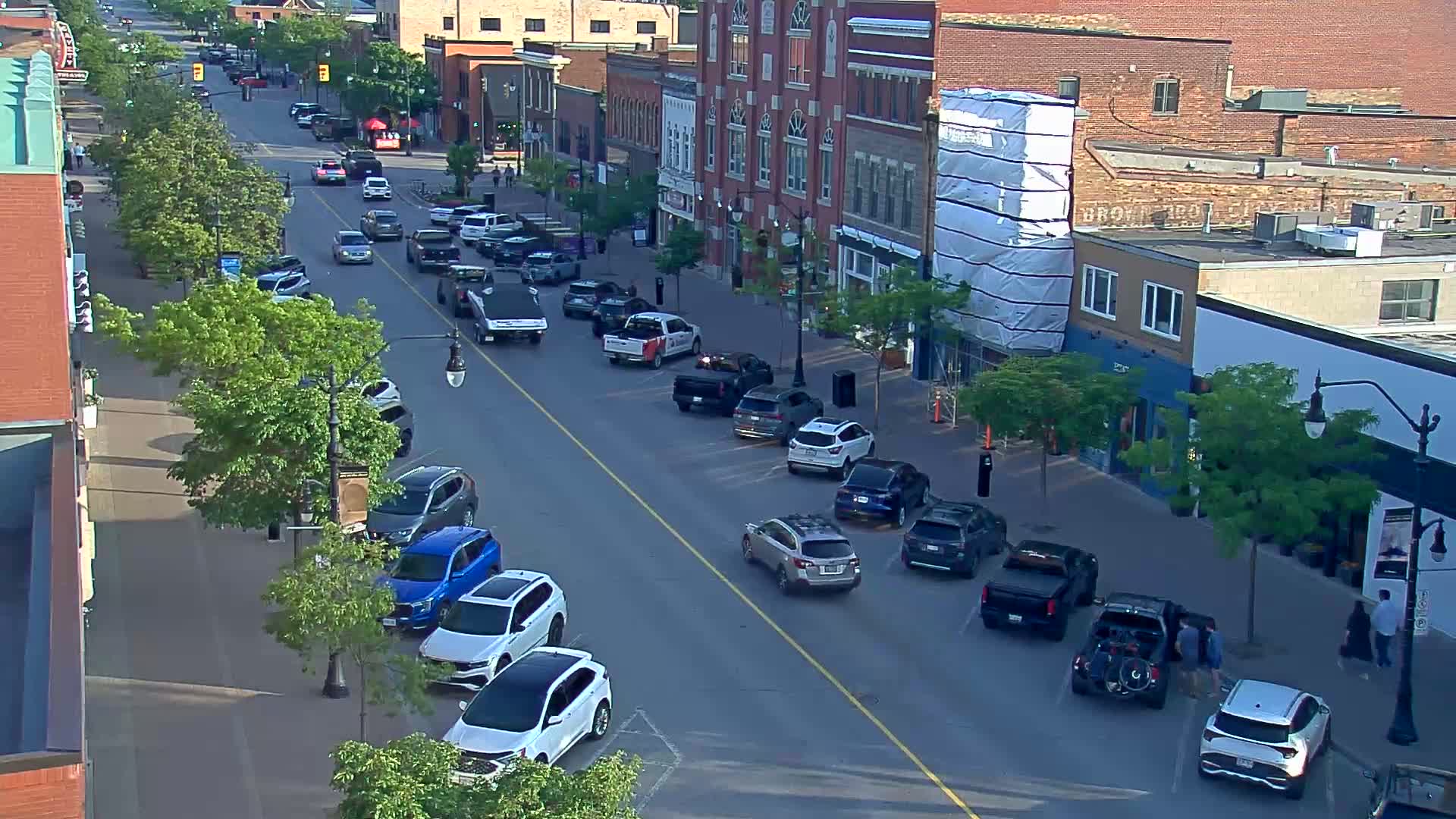 A sunny street scene shows cars parked along both sides of a road in front of brick buildings, with pedestrians walking on the sidewalk and trees lining the street.