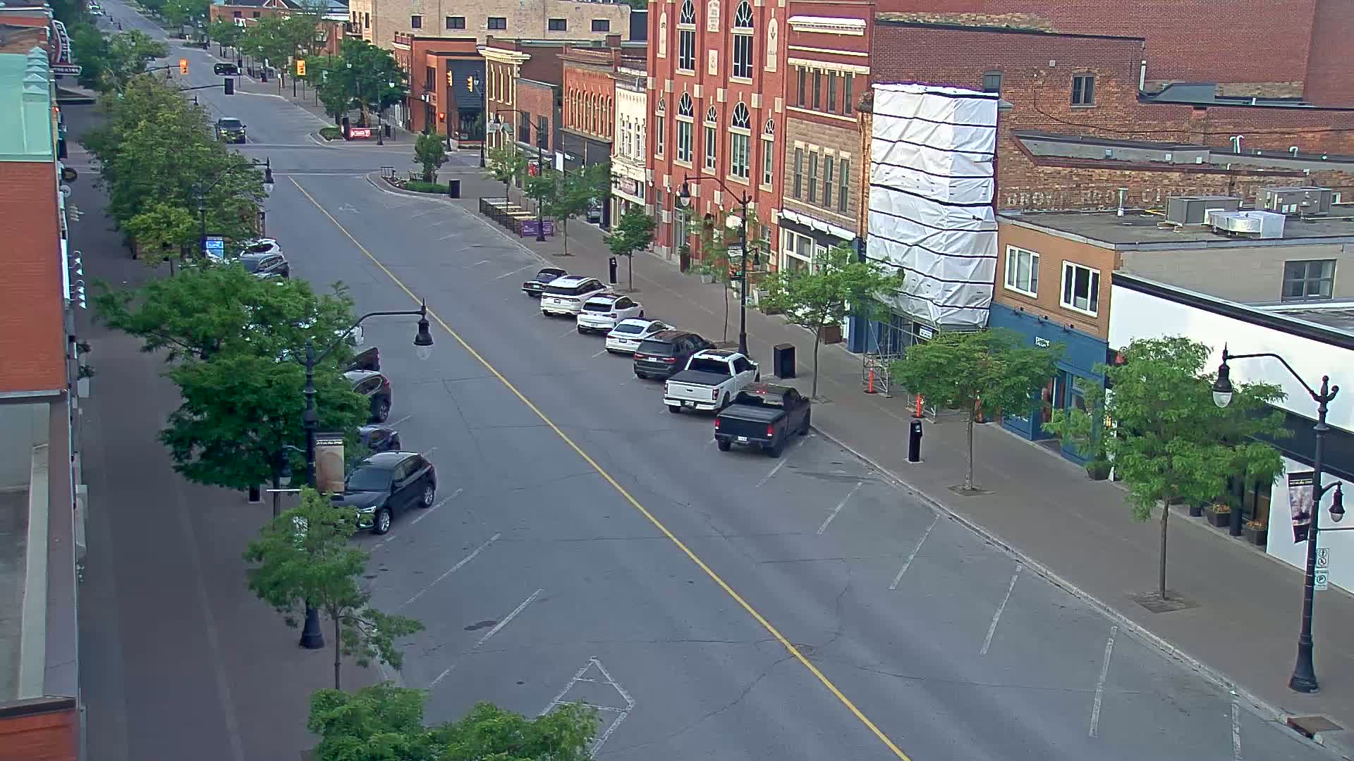 A mostly empty street lined with parked cars and brick buildings, some with trees in front, on a partly cloudy day.
