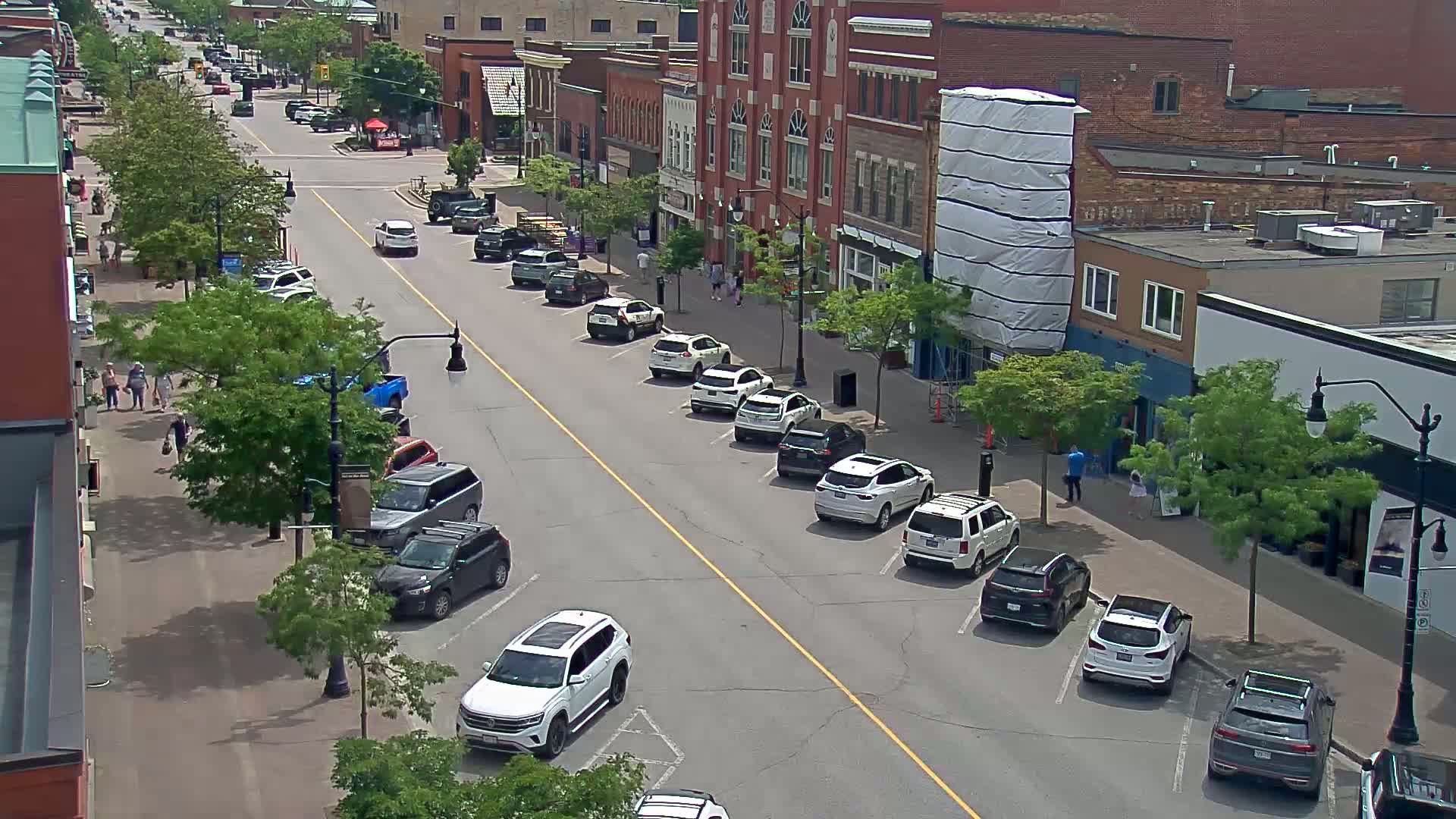 A sunny street scene shows cars parked on both sides of a city street lined with brick buildings and leafy trees, with pedestrians walking on the sidewalk.