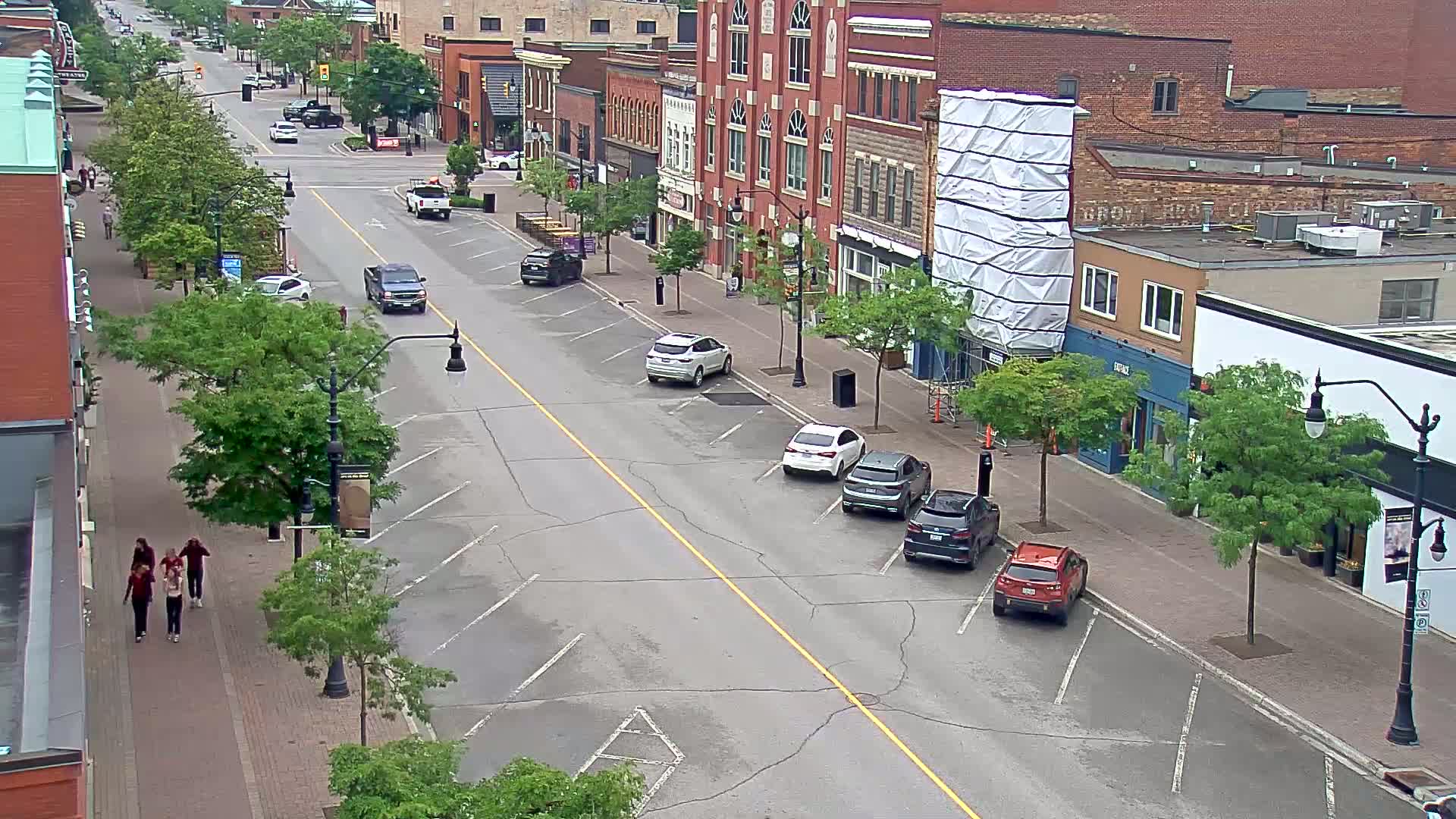 A mostly empty street lined with parked cars and brick buildings on a cloudy day, with a few pedestrians walking on the sidewalk.