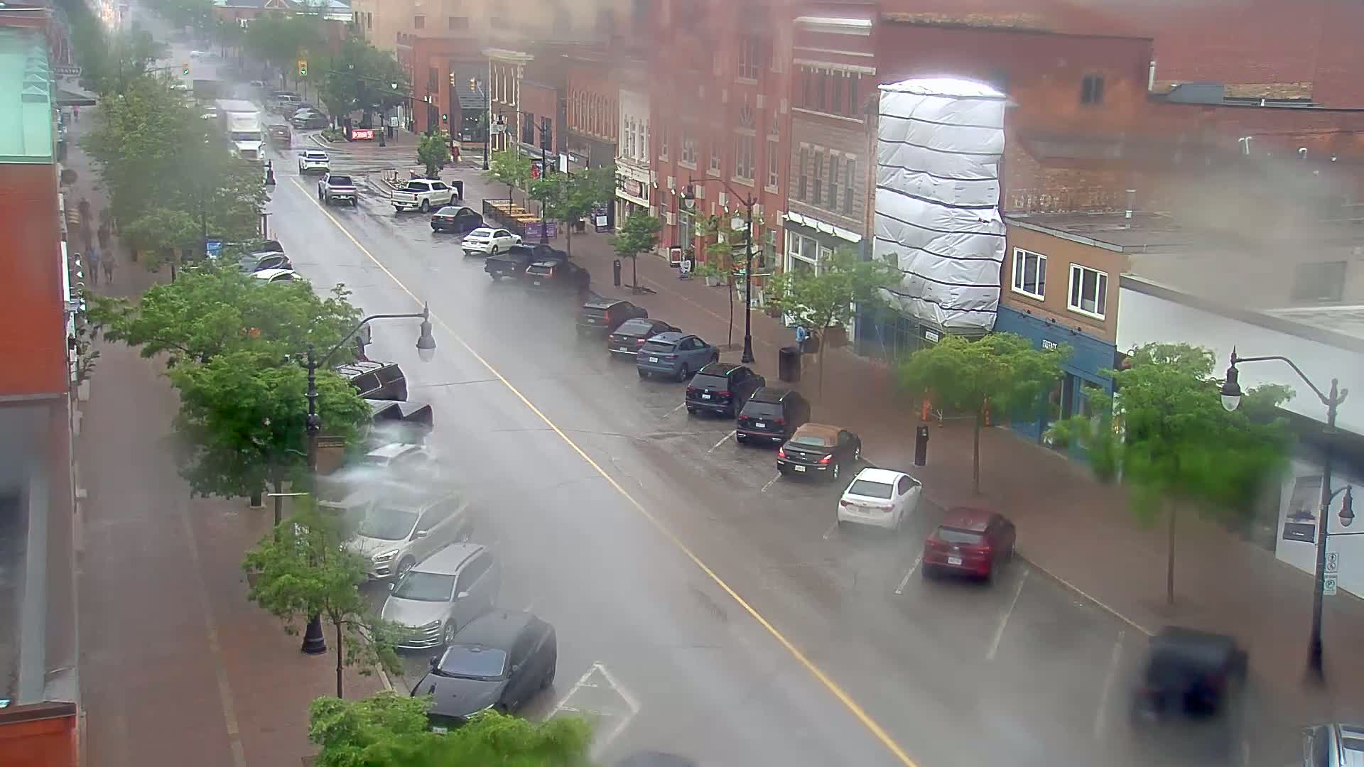 A rain-soaked city street lined with parked cars and buildings, some of which are under construction.