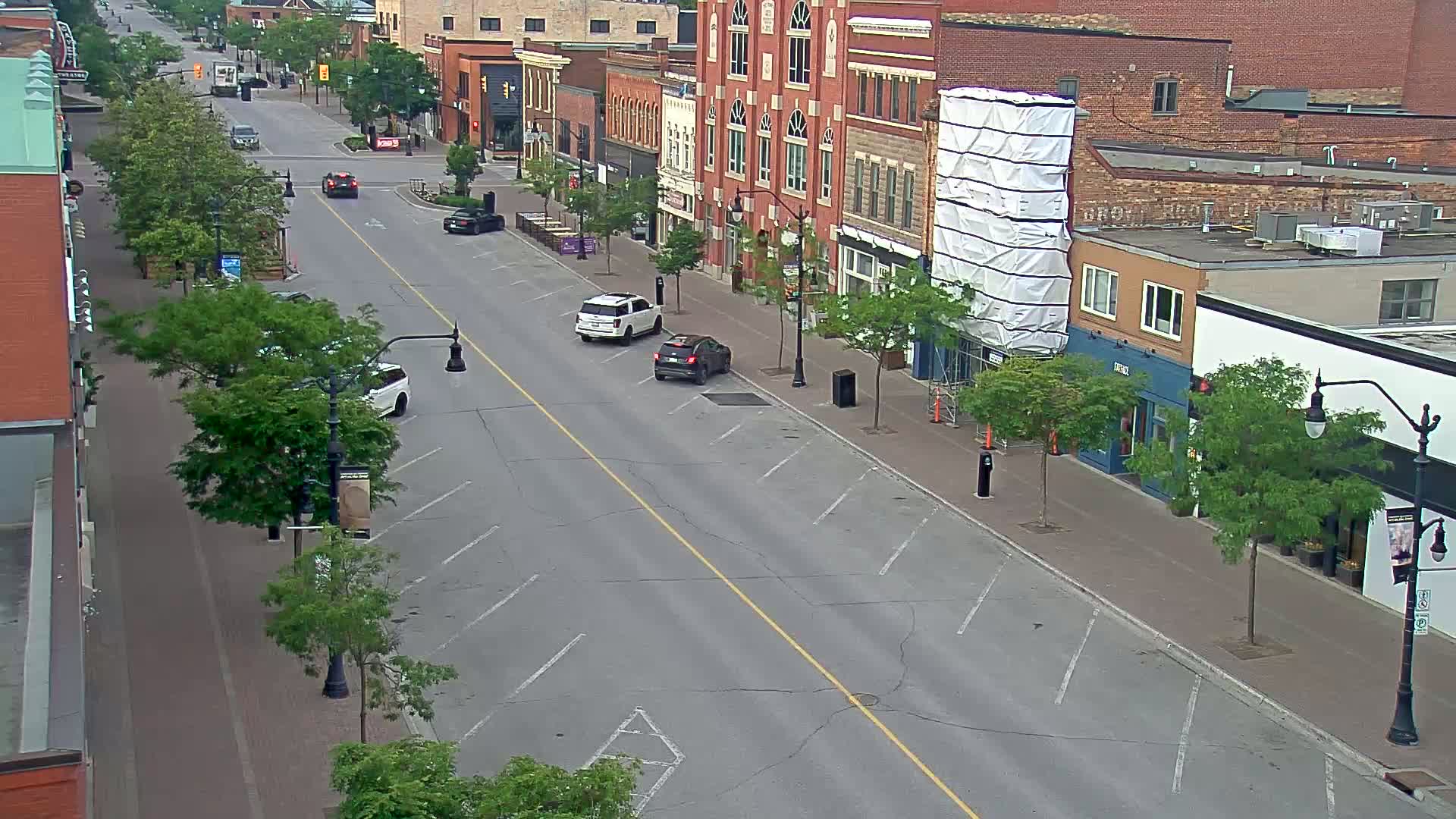A mostly empty street lined with brick buildings and leafy trees on a partly sunny day, with a few cars parked along the sides.