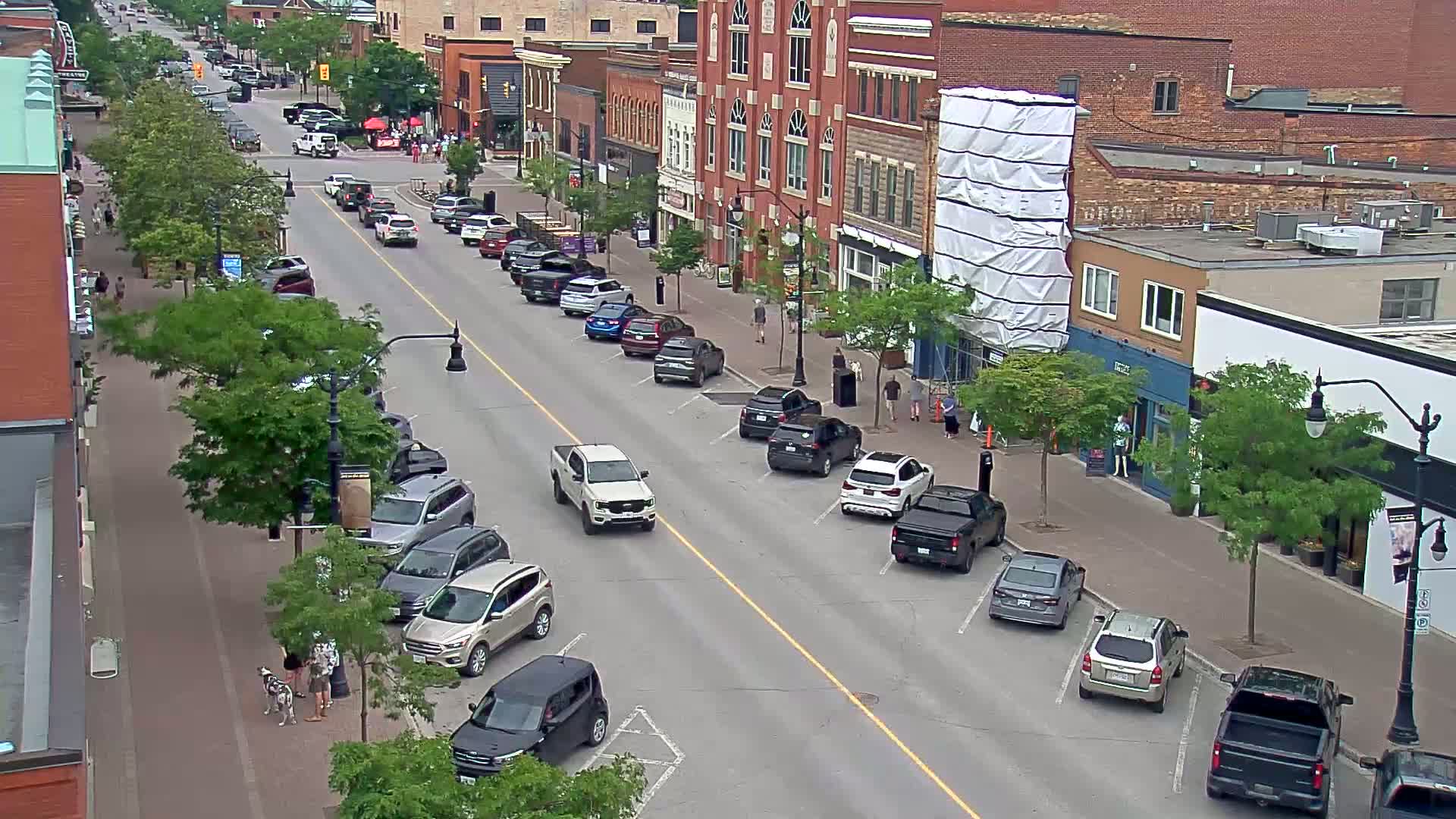 A street scene on a sunny day shows cars parked on both sides of a city street, in front of brick buildings, with pedestrians walking on the sidewalks.