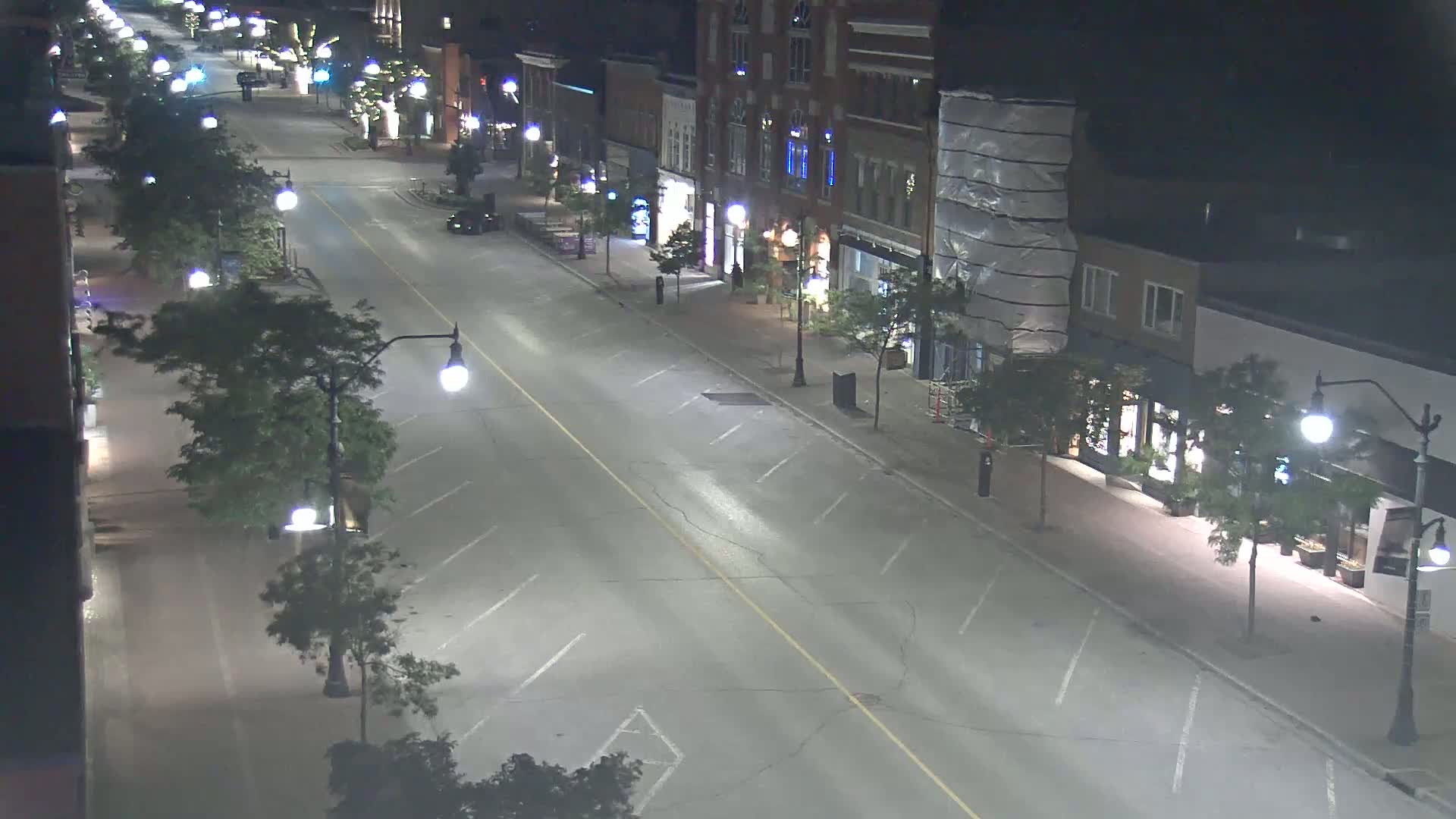 A mostly empty, brightly lit street at night, lined with two-story buildings and trees, shows a few parked cars.