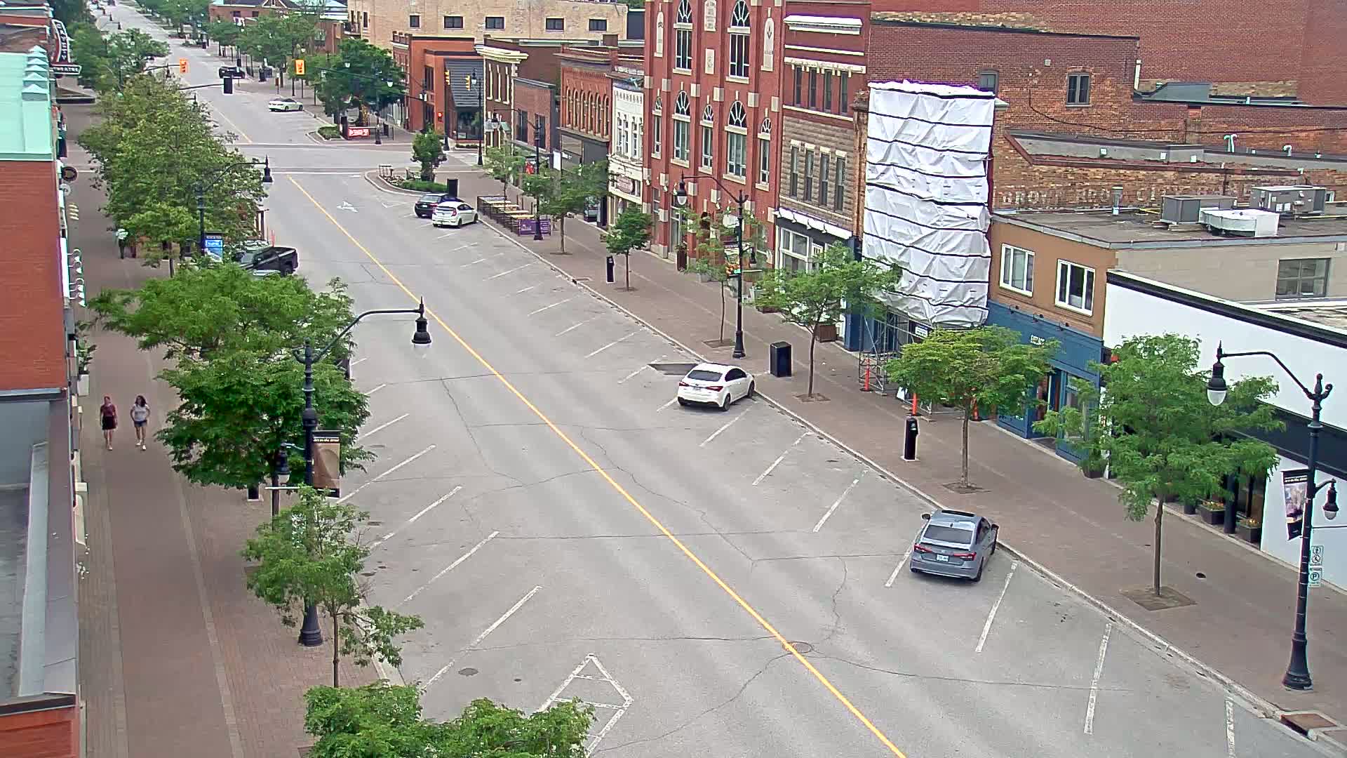 A mostly empty street lined with brick buildings and parked cars on a partly sunny day, with two pedestrians walking on the sidewalk.