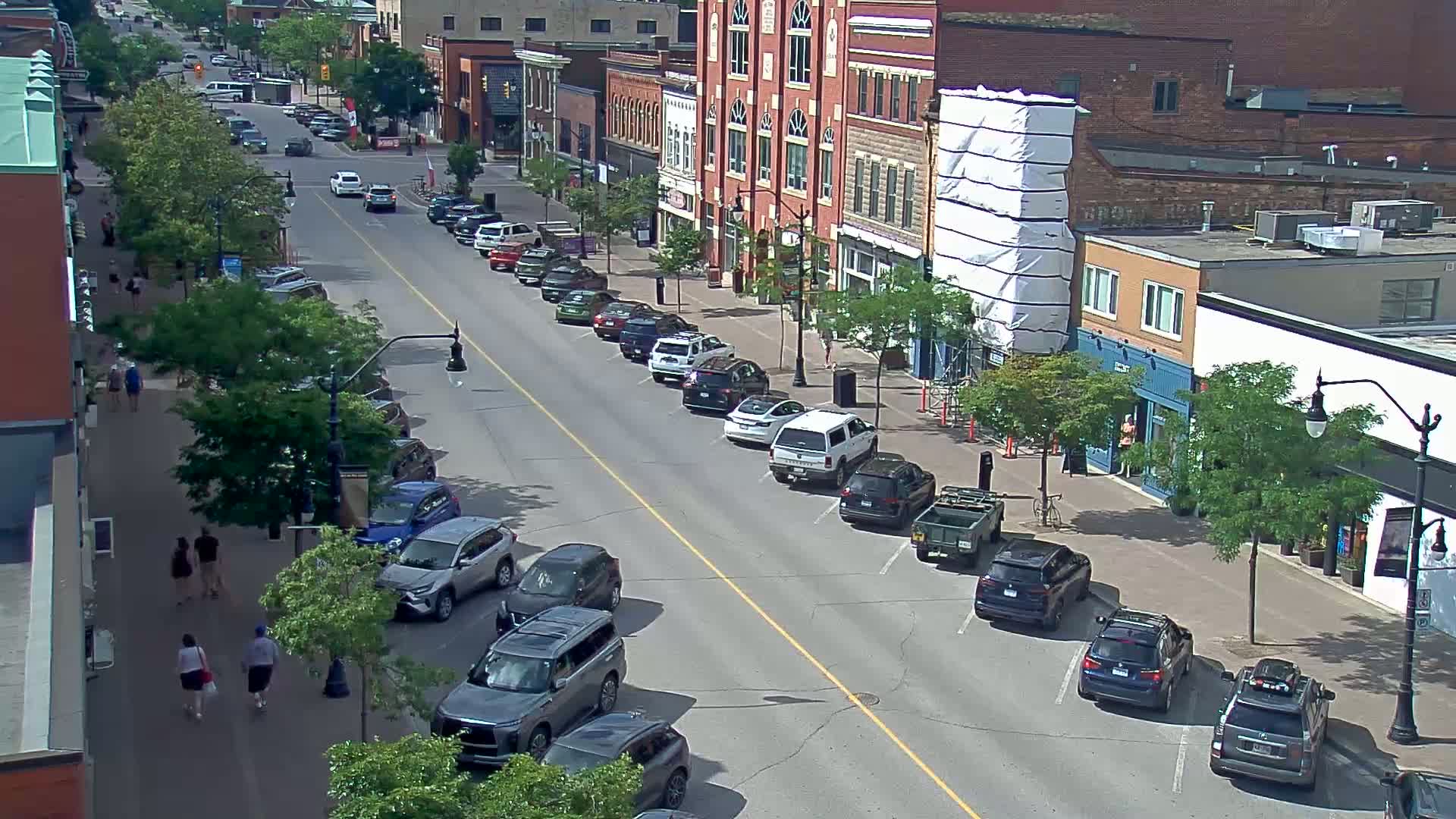 A sunny day on a city street lined with parked cars and pedestrians walking past brick buildings, one of which is under construction.