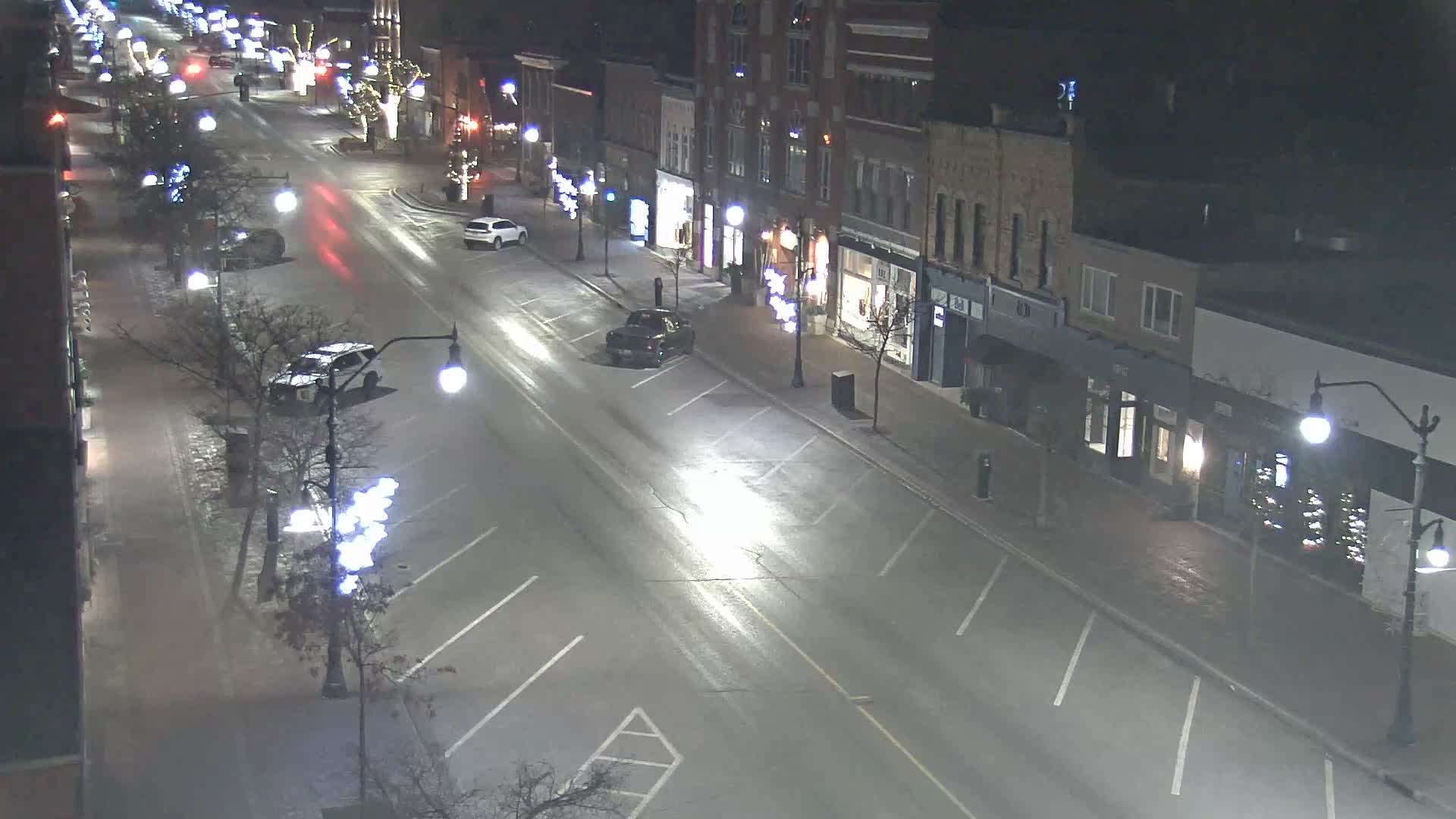 An elevated nighttime view shows a quiet street lined with multi-story buildings and bare trees, illuminated by streetlights and festive white decorations, with several cars on wet pavement.
