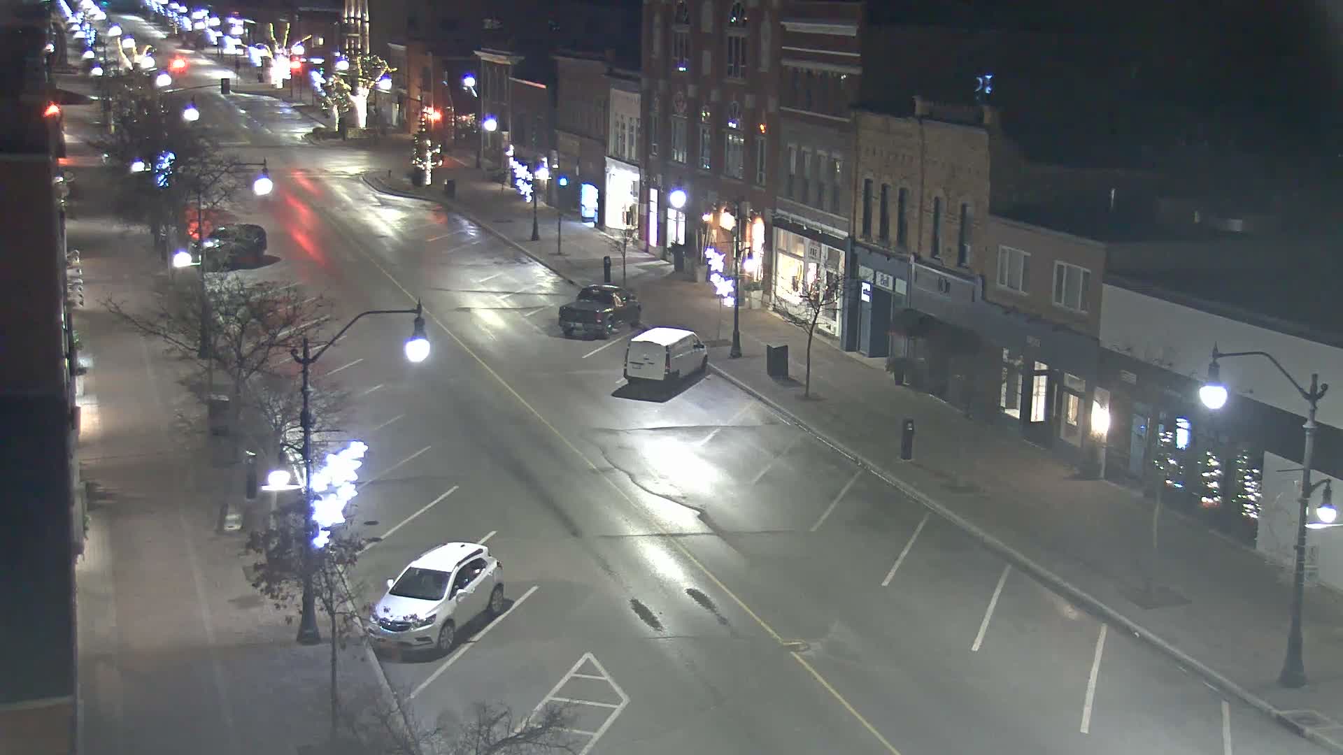 An elevated view captures a well-lit downtown street at night, featuring historic buildings with glowing storefronts, bare trees adorned with white lights, and several parked vehicles, all on a clear and wet night.