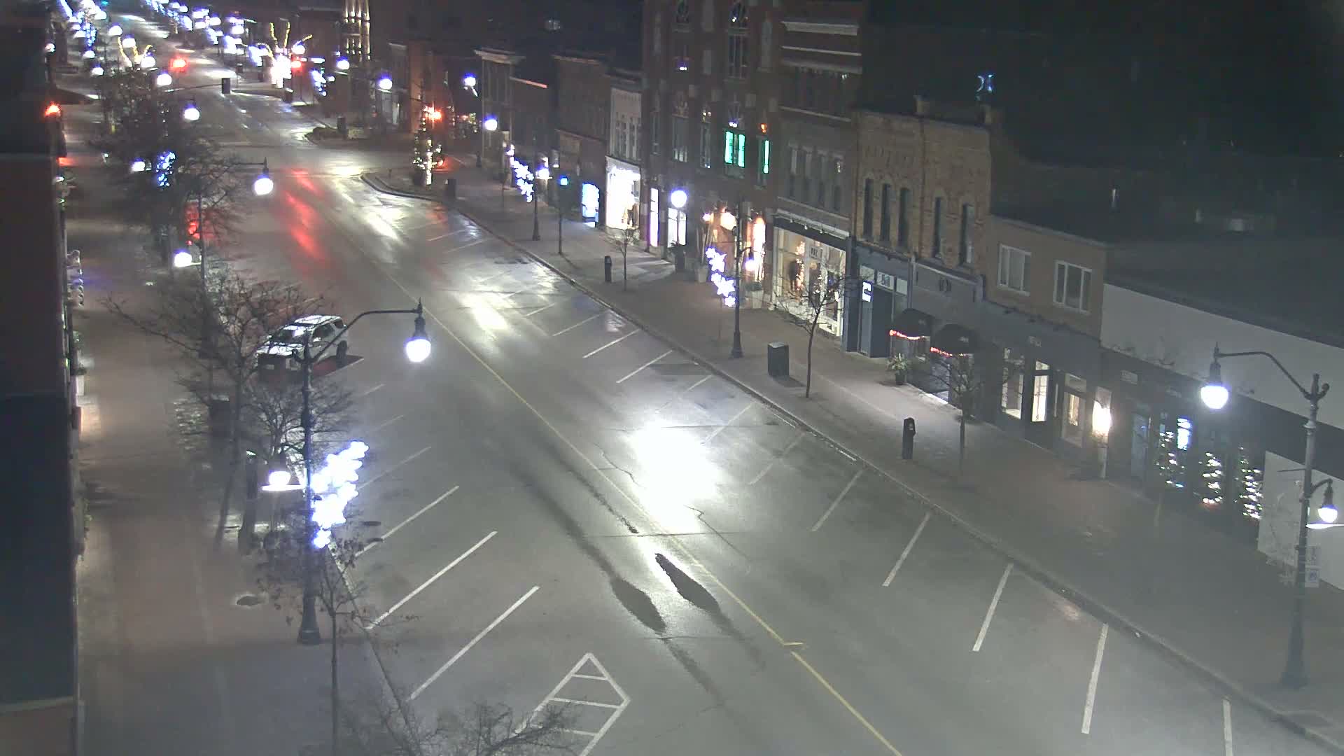 An elevated nighttime view captures a wet urban street with sparse traffic, lined by brick buildings, bare trees adorned with decorative lights, and bright streetlights reflecting on the damp pavement.