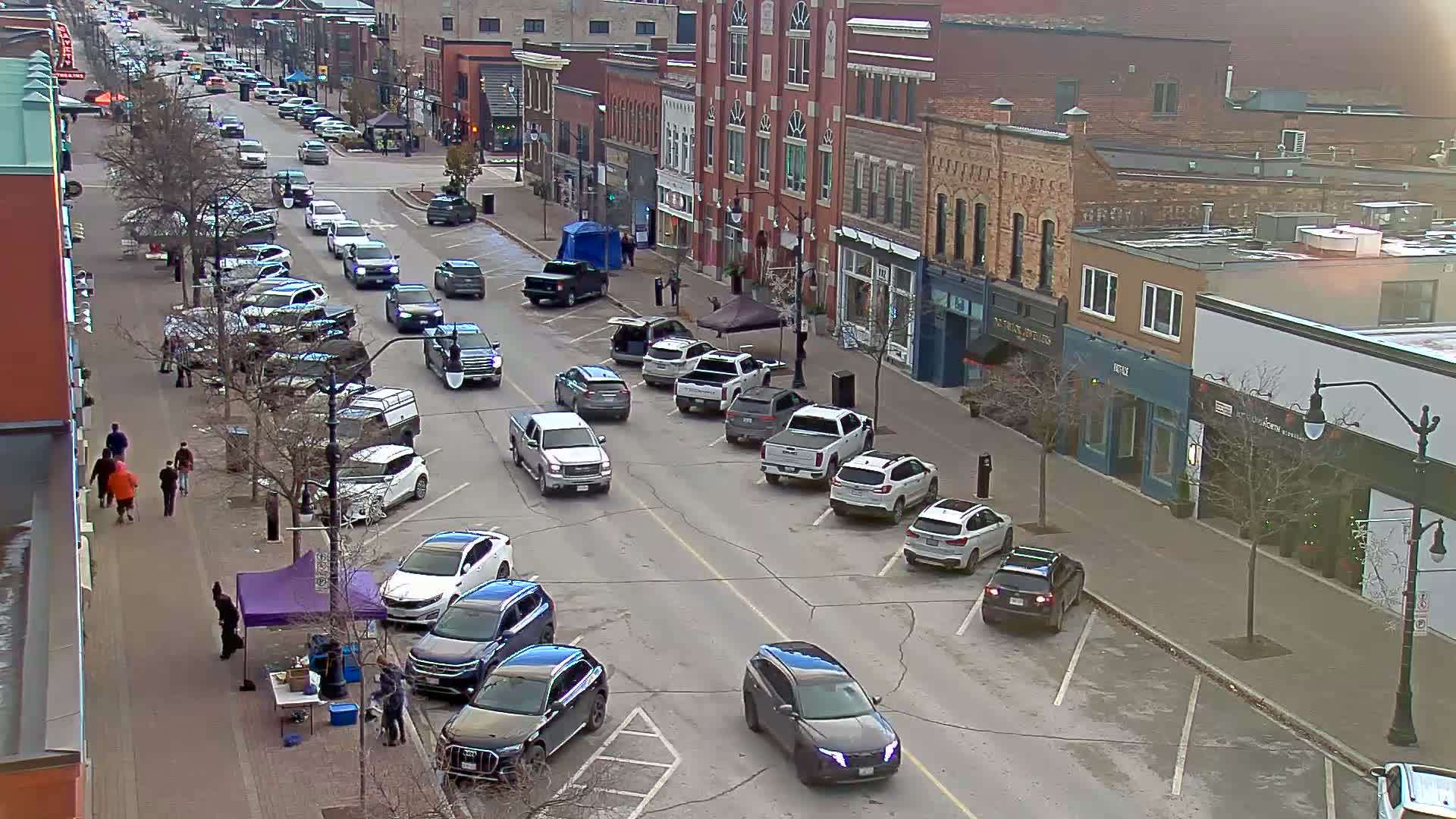 A bustling downtown street is seen from an elevated perspective, featuring numerous parked and moving vehicles, pedestrians on sidewalks, bare trees, and temporary market tents, all under cool, overcast daylight.