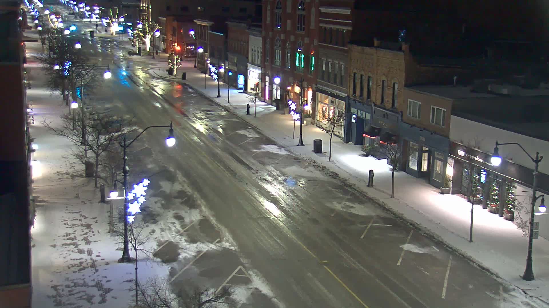 A quiet city street is seen at night under snowy and wet conditions, with buildings illuminated by streetlights and festive holiday decorations lining the sidewalks.