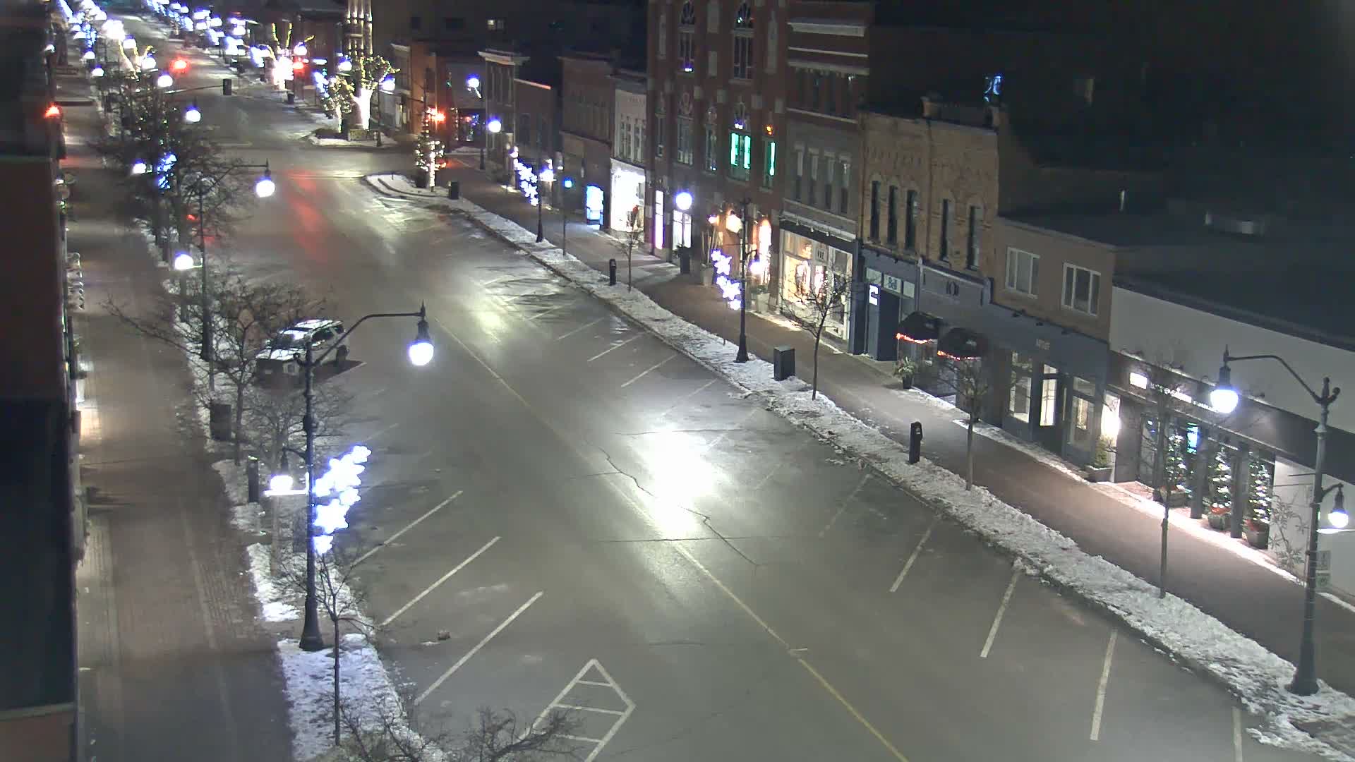 A quiet, snow-dusted downtown street, lined with buildings illuminated by streetlights and festive decorations, is seen under a clear, cold night sky with a solitary car parked on the left.