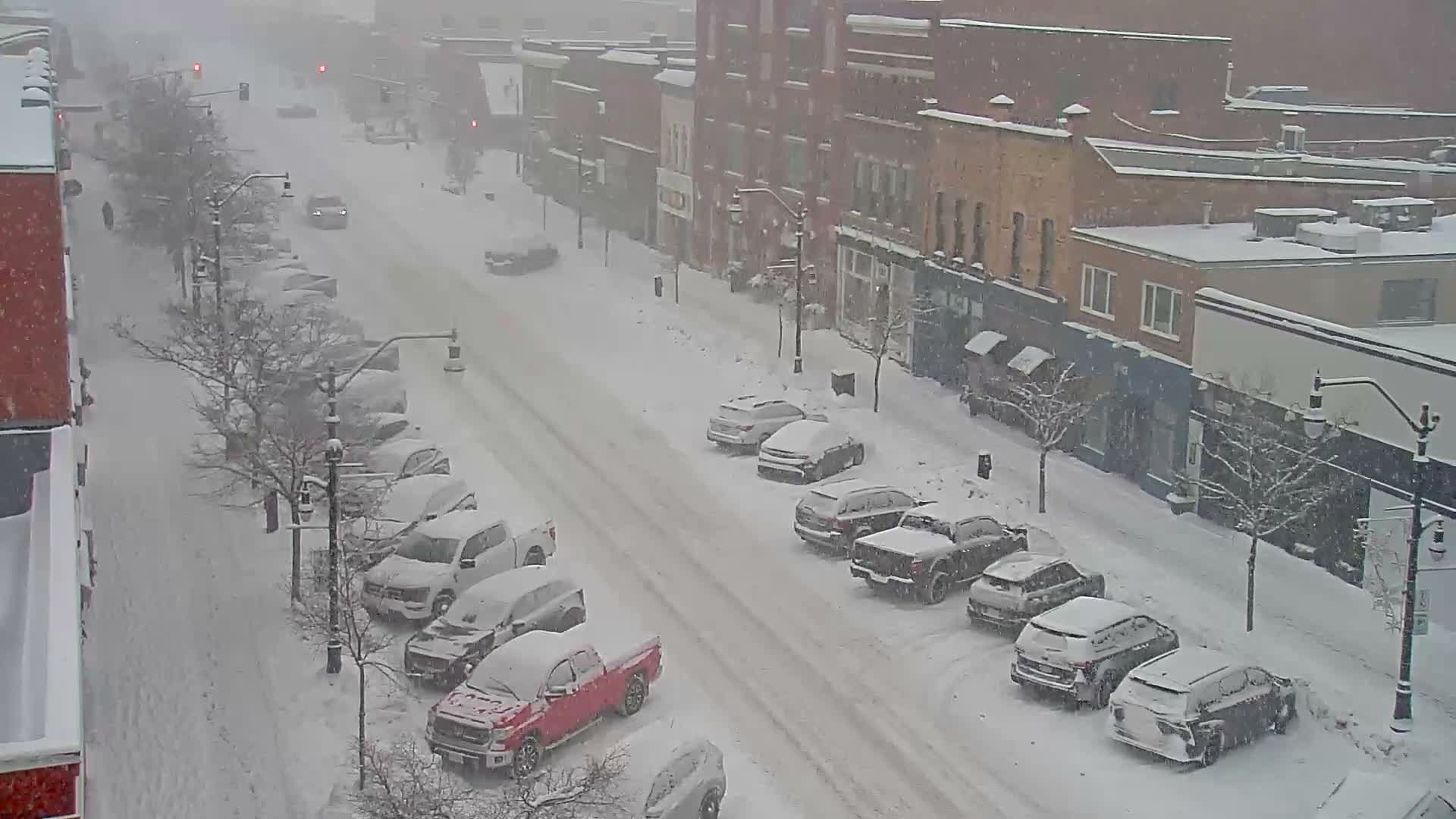 A quiet, snow-dusted downtown street, lined with buildings illuminated by streetlights and festive decorations, is seen under a clear, cold night sky with a solitary car parked on the left.