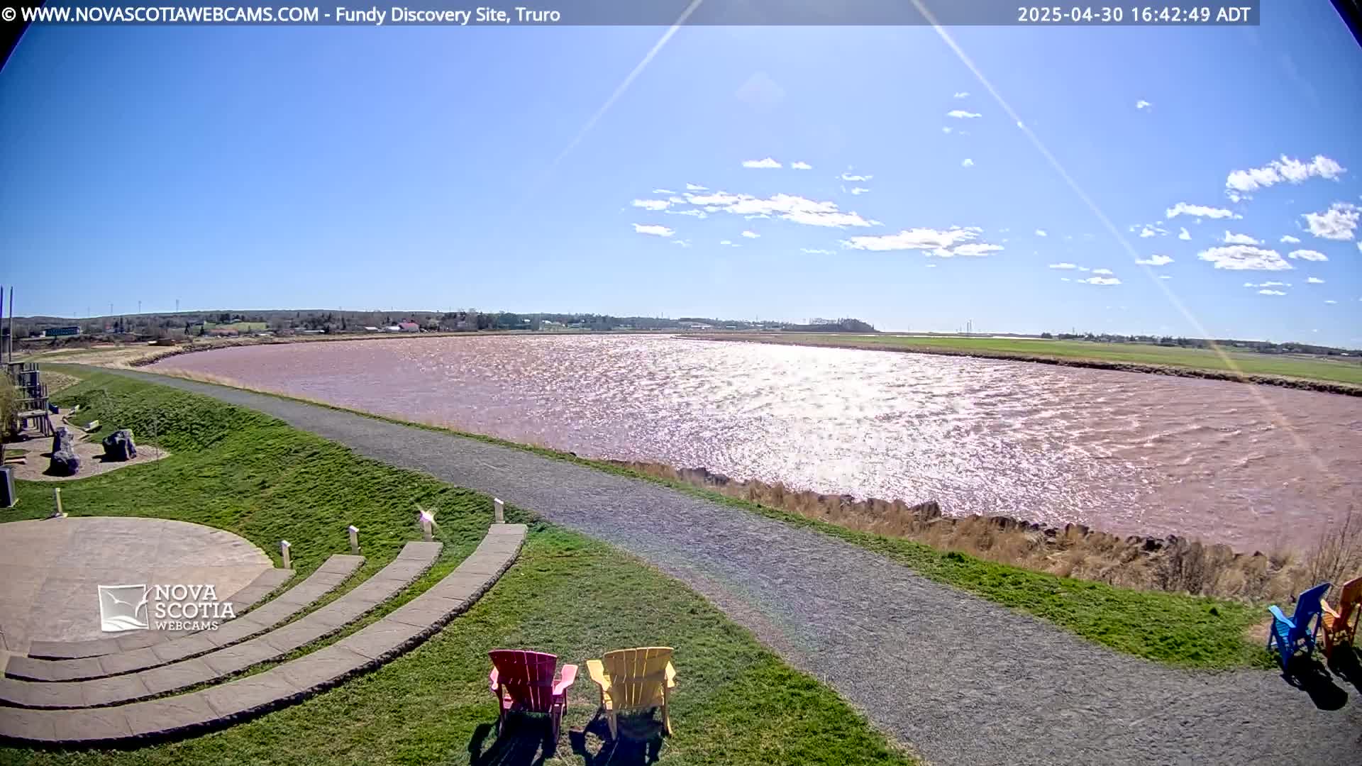 A pinkish-red body of water is seen under a mostly sunny sky, with a paved path and colorful Adirondack chairs situated on a grassy area overlooking it.