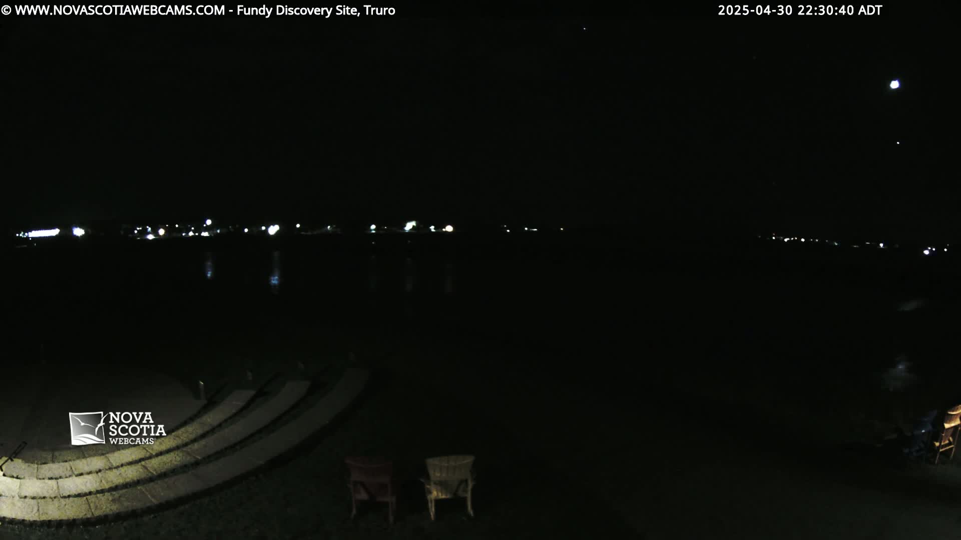 A nighttime view of a dark body of water with distant lights on the horizon, a moon in the sky, and empty chairs on a stone amphitheater in the foreground.