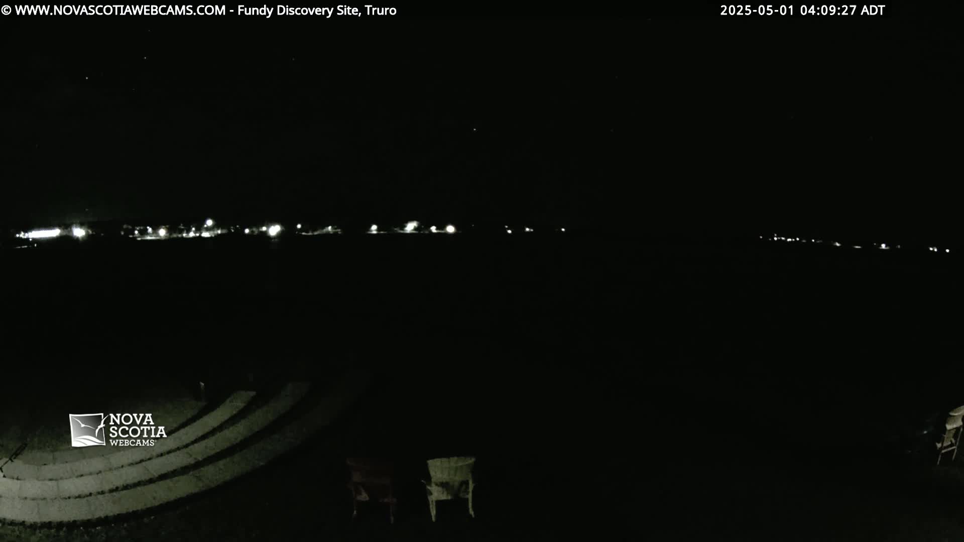 A nighttime view of a distant line of lights, a dark amphitheater-like structure, and a couple of chairs in the foreground under a clear, dark sky.