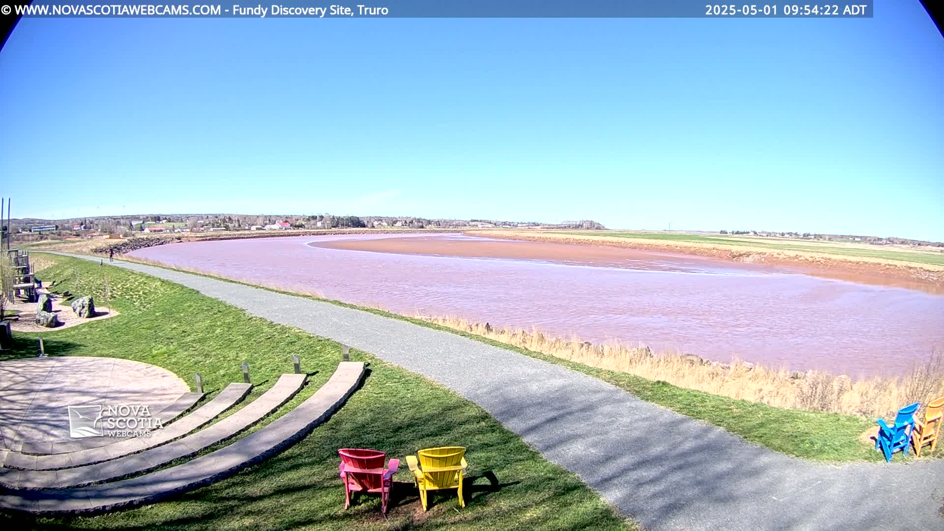 A paved walkway leads to a pinkish-red river under a clear blue sky, with grassy areas, a stone amphitheater, and a few colorful chairs visible in the foreground.