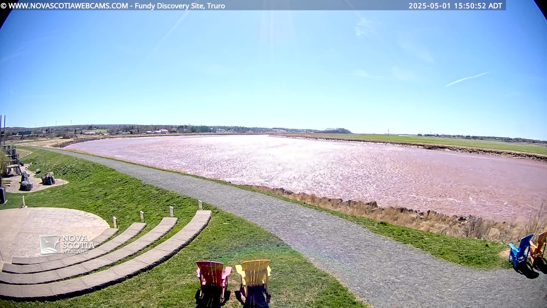 A pinkish-red river stretches under a clear blue sky, viewed from a grassy area with a paved path and a few Adirondack chairs.