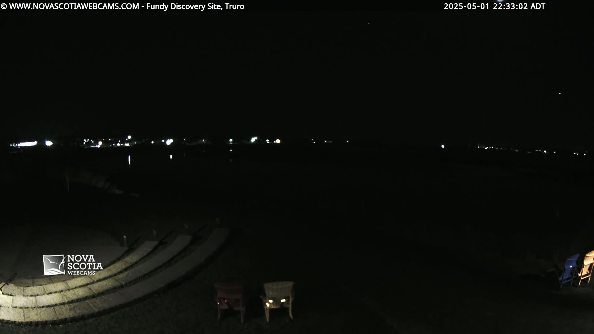 A nighttime view of a dark, empty amphitheater overlooking a calm body of water with distant city lights under a clear sky.