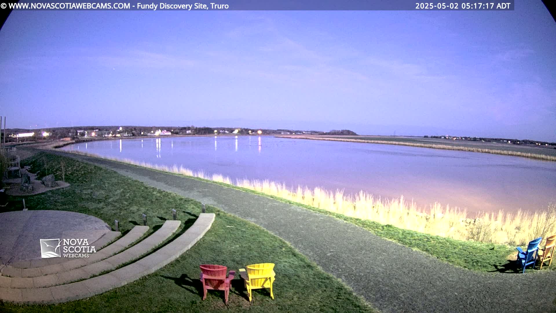 A calm, twilight-lit body of water stretches out before a grassy area with a stone amphitheater and three brightly colored Adirondack chairs.