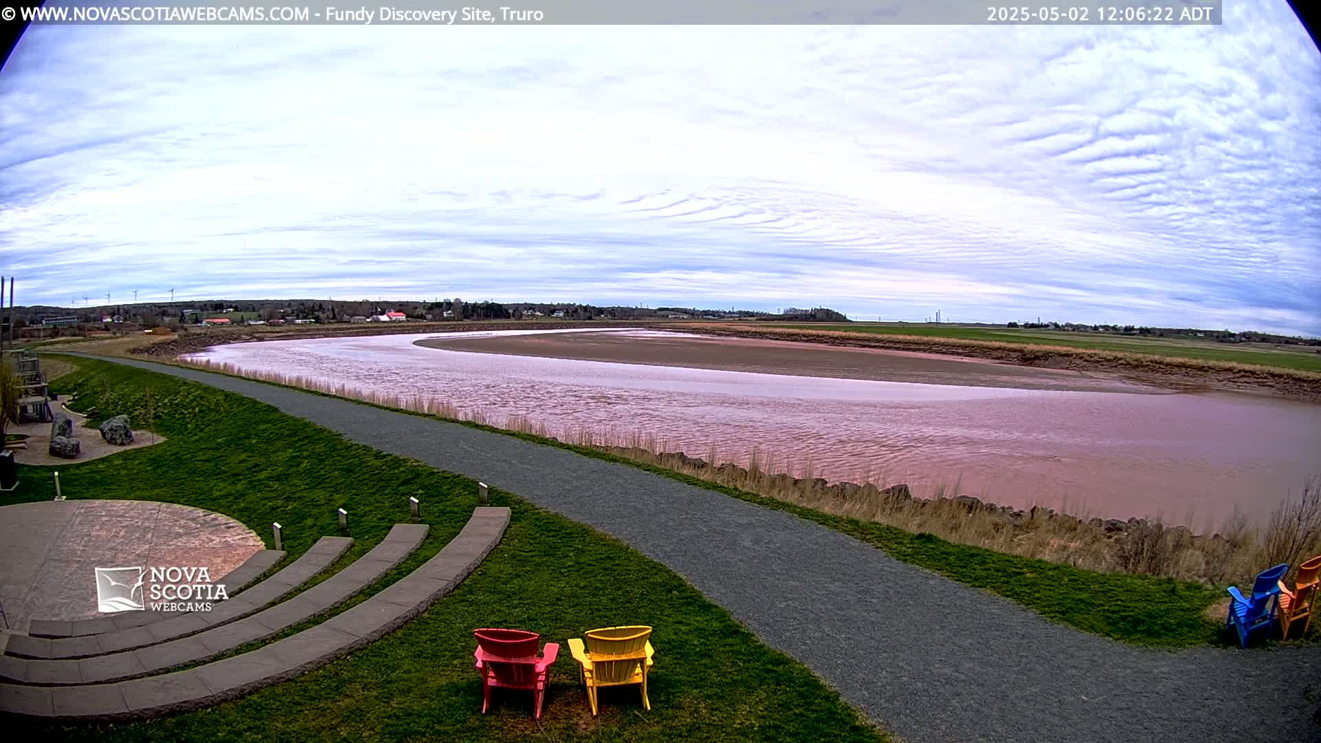 A wide, pinkish-red river curves gently through a grassy landscape under a mostly cloudy sky, with three Adirondack chairs positioned near a paved pathway overlooking the water.