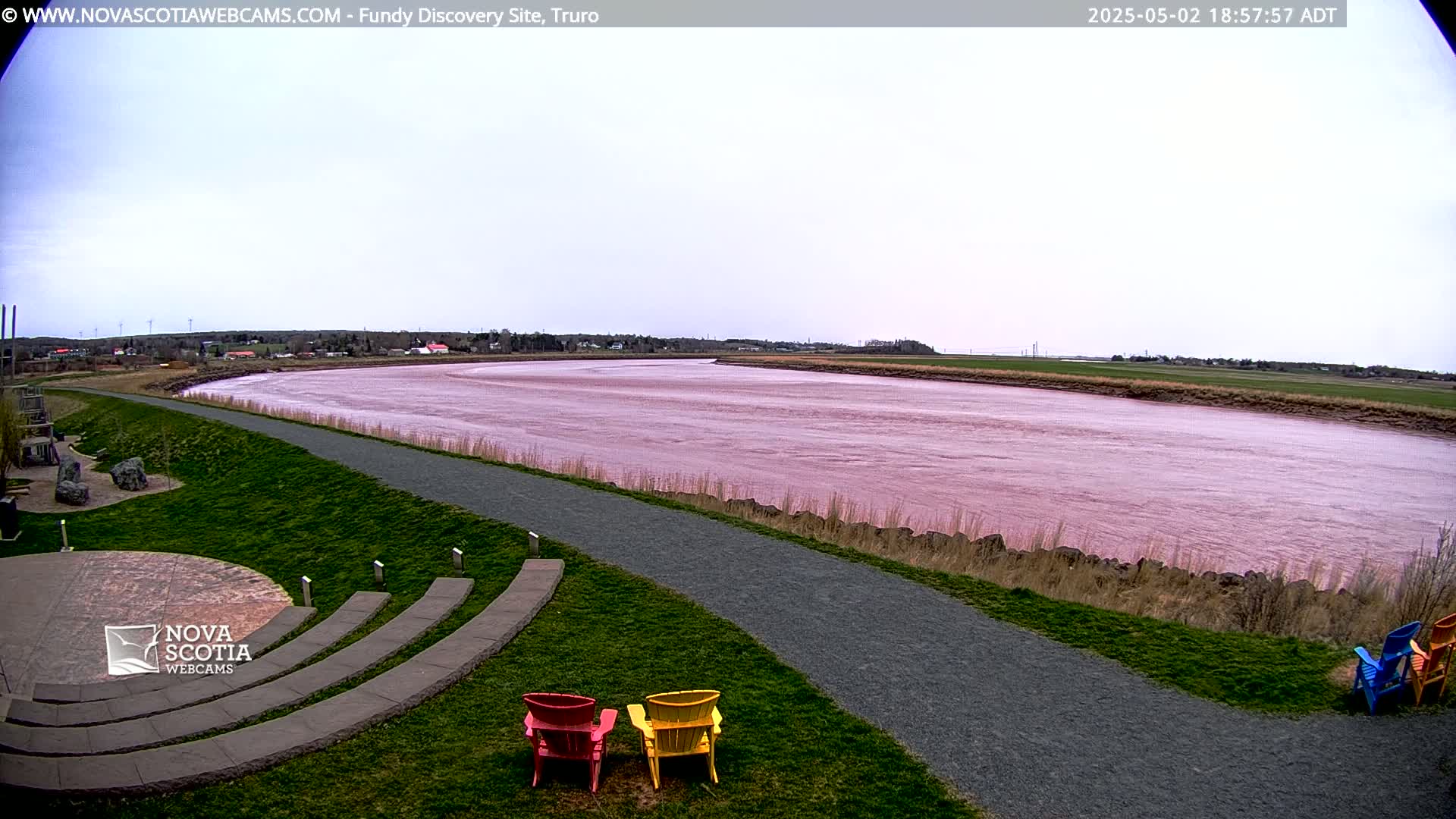 A pinkish-red river flows under an overcast sky, beside a grassy area with a paved path and several colorful Adirondack chairs.