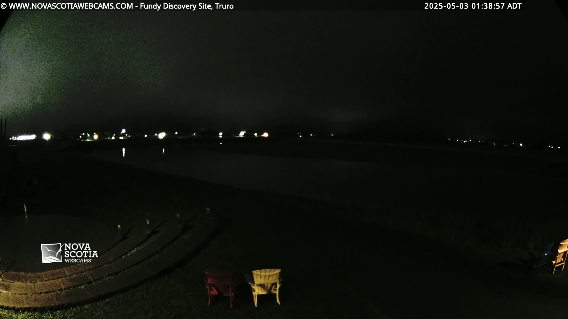 A dark nighttime scene shows a body of water with distant lights, an empty amphitheater, and two Adirondack chairs in the foreground under a cloudy sky.