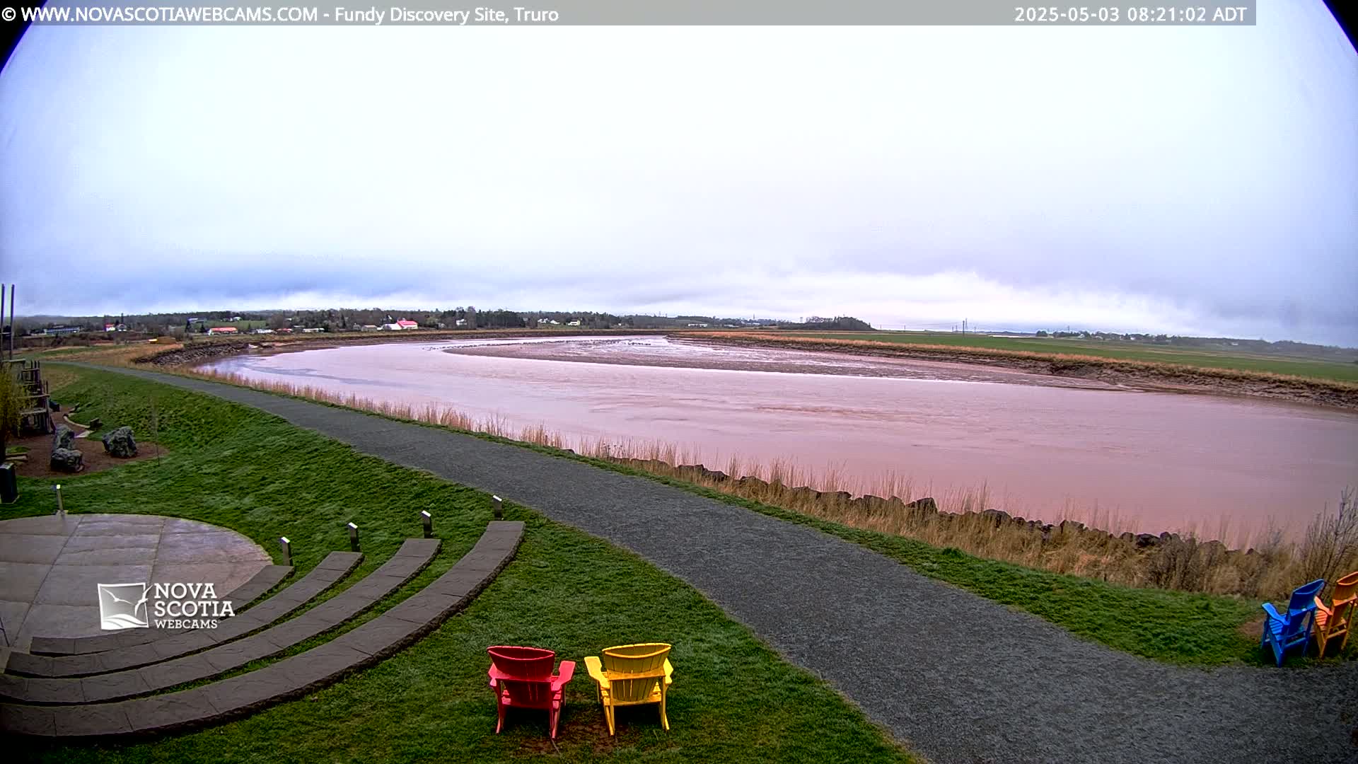 A wide, pinkish-brown river winds through a flat landscape under a cloudy sky, with a paved walkway and several colorful Adirondack chairs in the foreground.