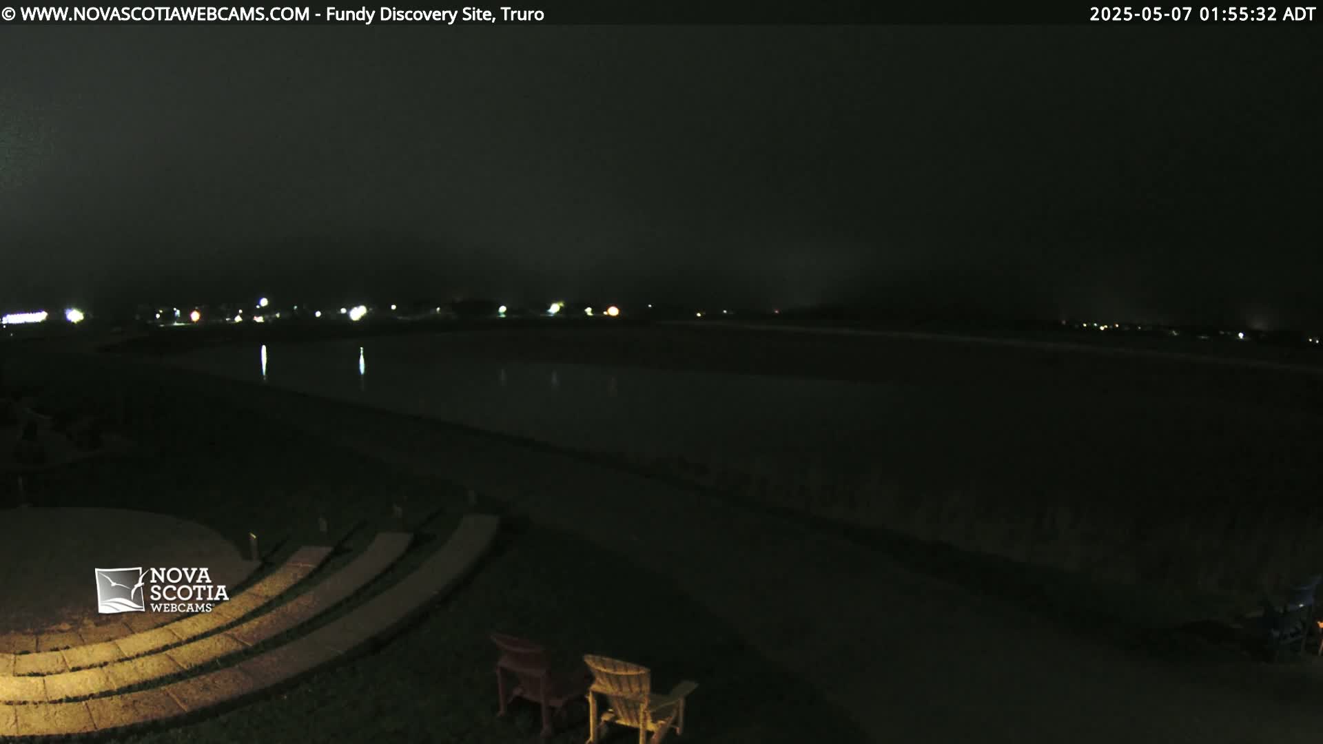 A nighttime view of a dark body of water, with dimly lit buildings in the distance and a stone amphitheater with two empty chairs in the foreground.