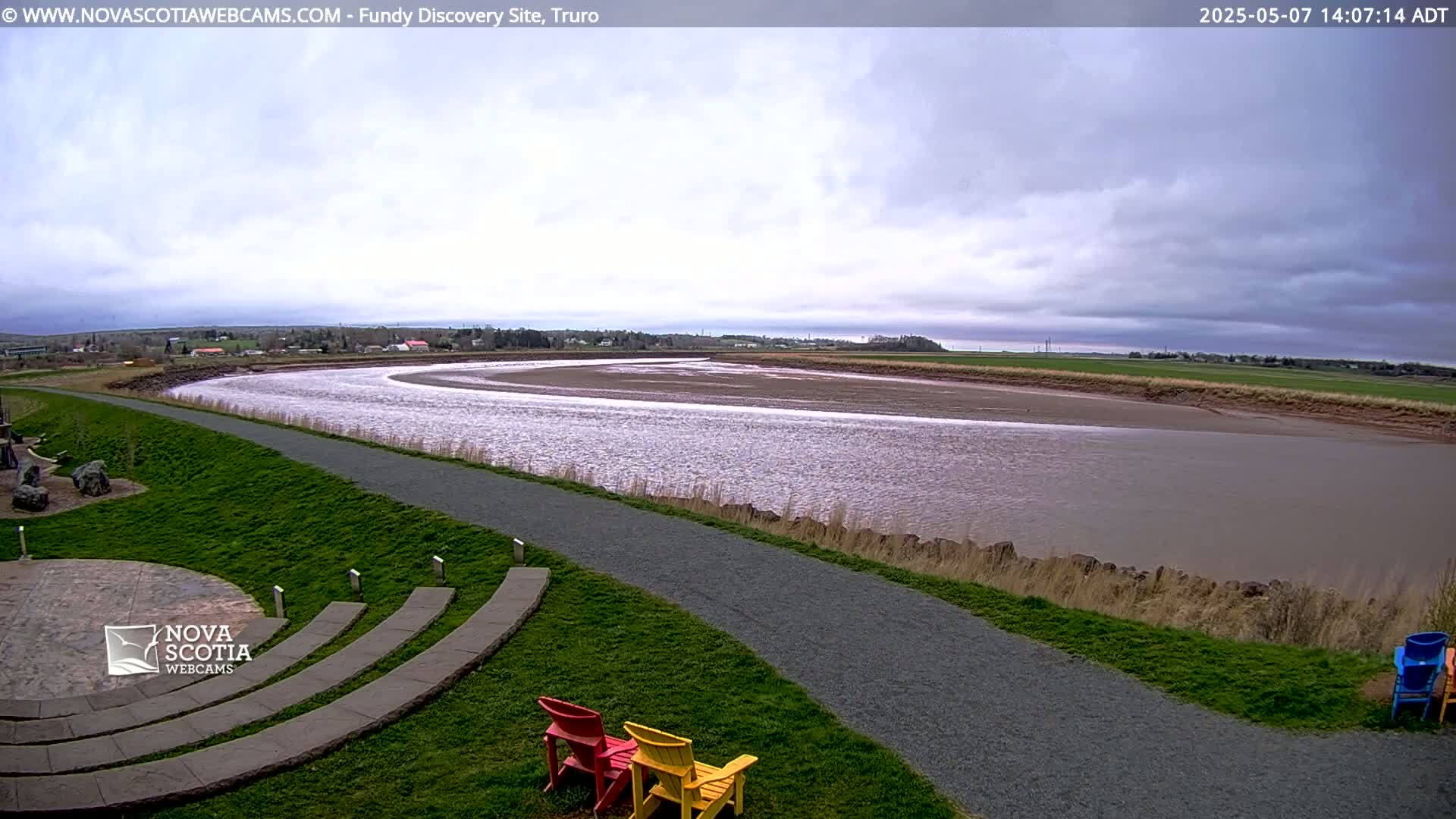 A wide, muddy river curves gently through a grassy landscape under a cloudy sky, with a paved path and outdoor seating nearby.