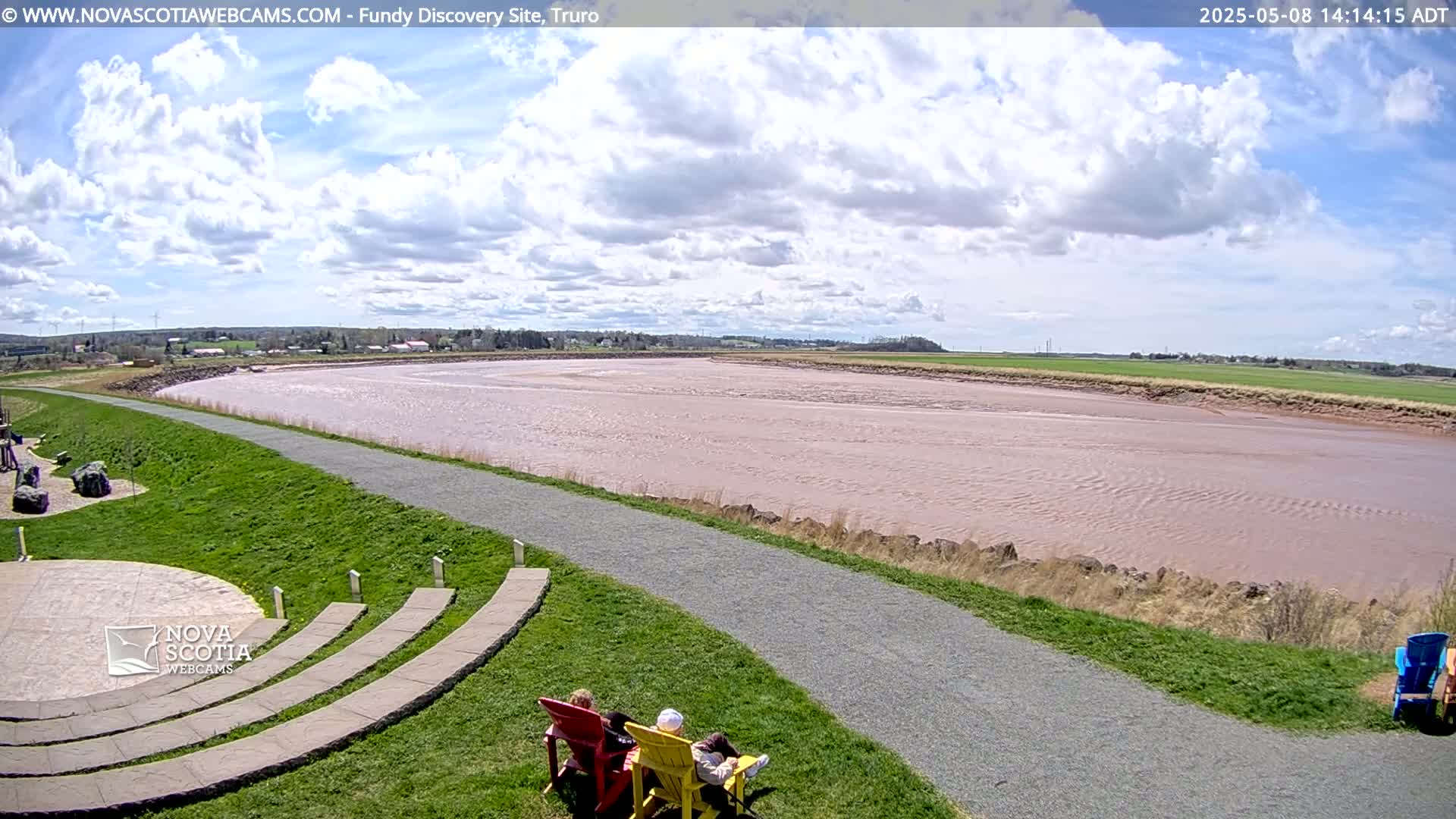A partly cloudy sky overlooks a wide, reddish-brown river, a grassy area with a paved path and stone seating, and a few people sitting in chairs.