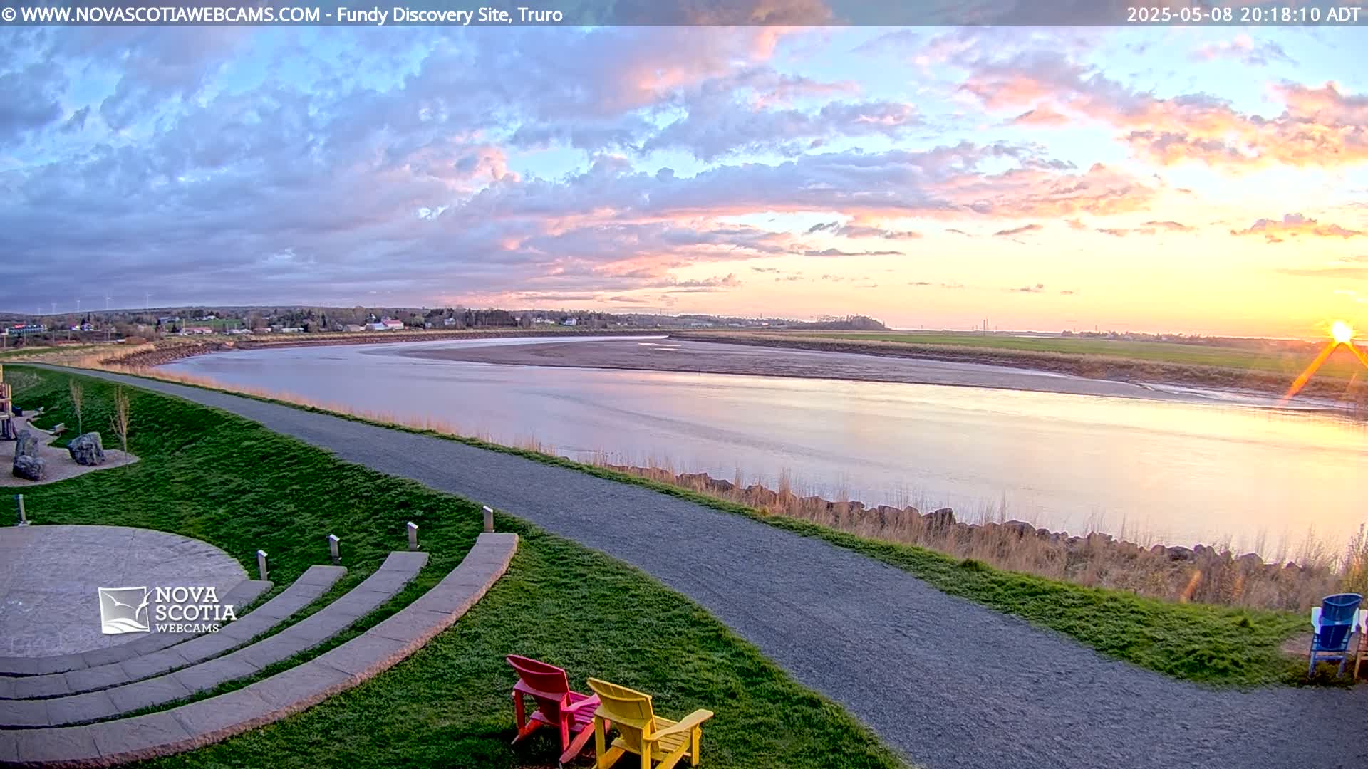 A wide river at sunset under a partly cloudy sky is viewed from a grassy area with a paved path and stone amphitheater seating.