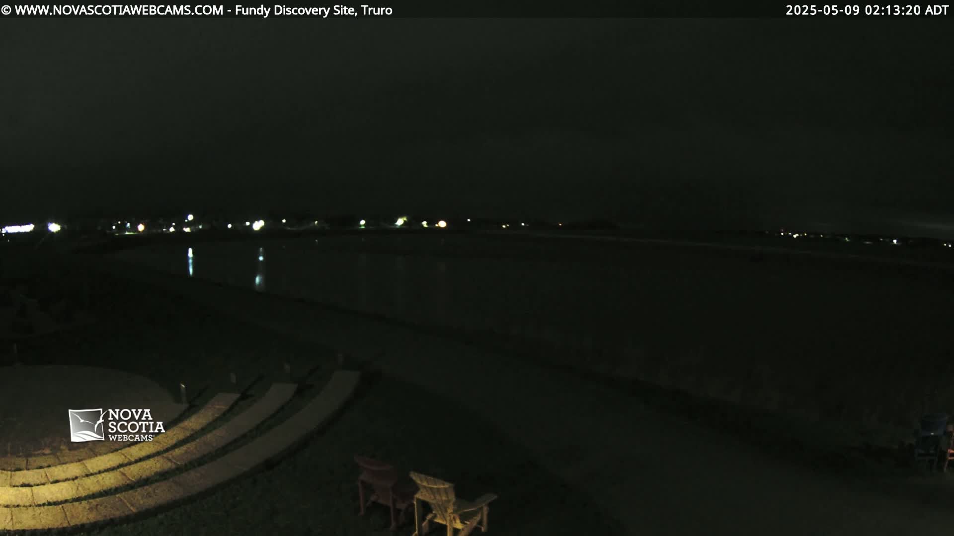 A nighttime view of a dark body of water reflecting distant lights, with empty chairs and a stone amphitheater in the foreground.