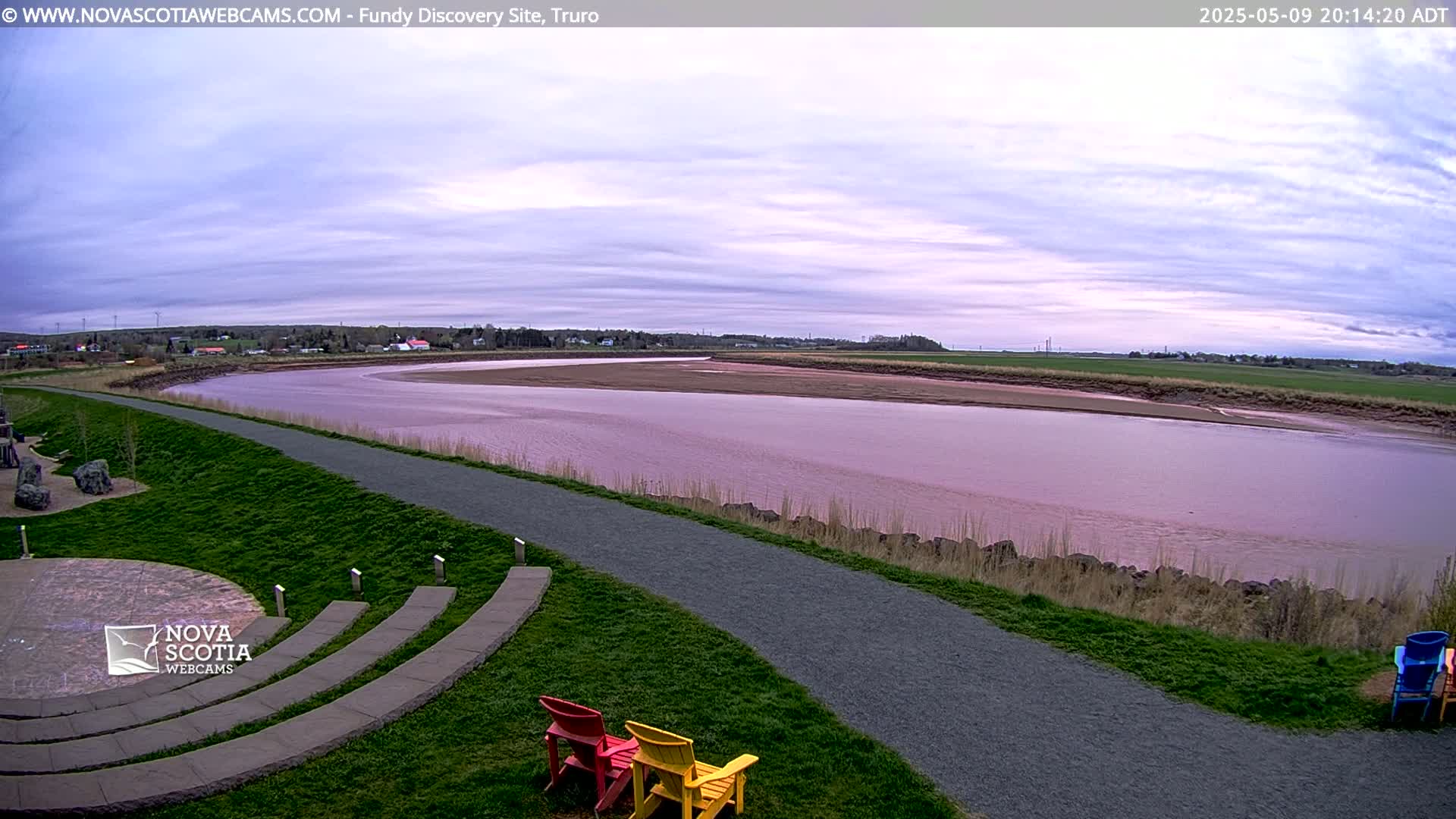 A wide, pinkish-brown river flows past a grassy area with a paved path and a few colorful chairs under an overcast sky.