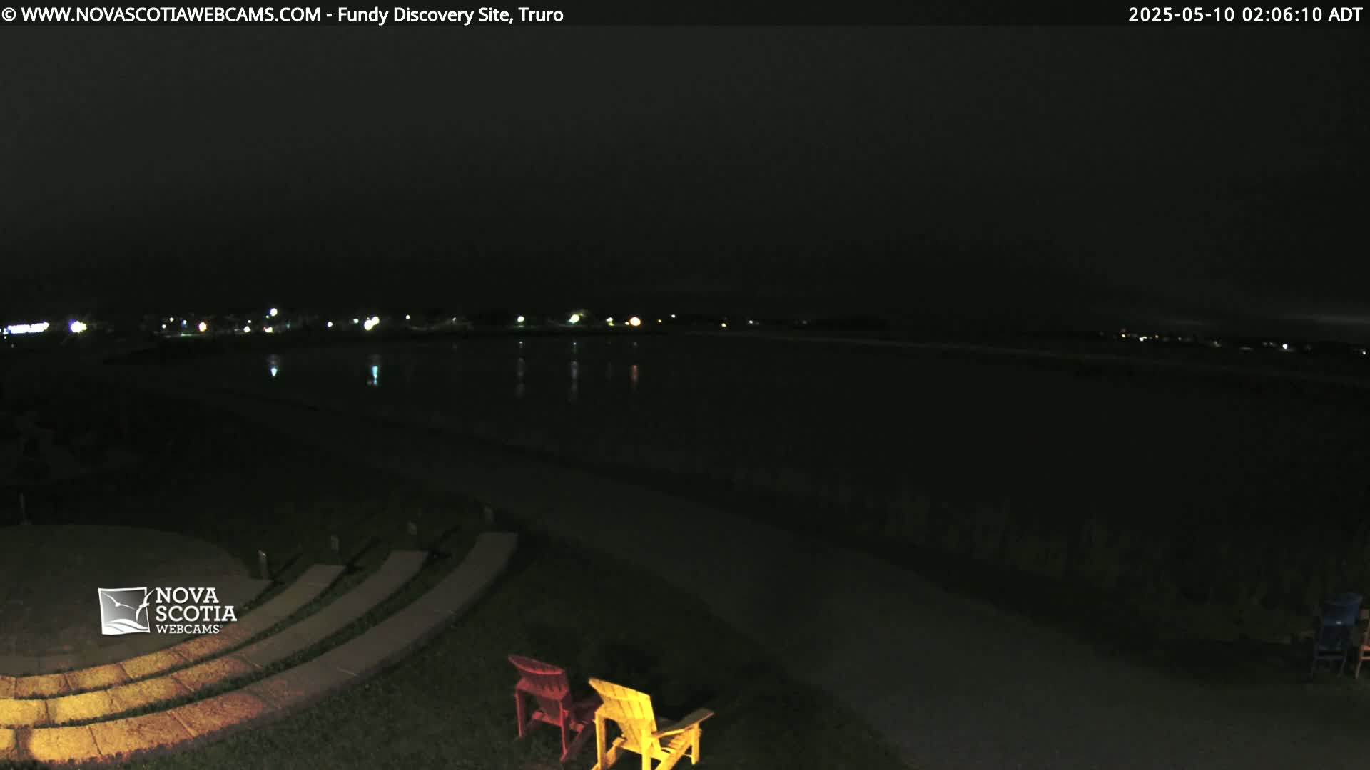 At night, two empty chairs sit near a stone amphitheater overlooking a dark body of water with faintly visible lights across the water.