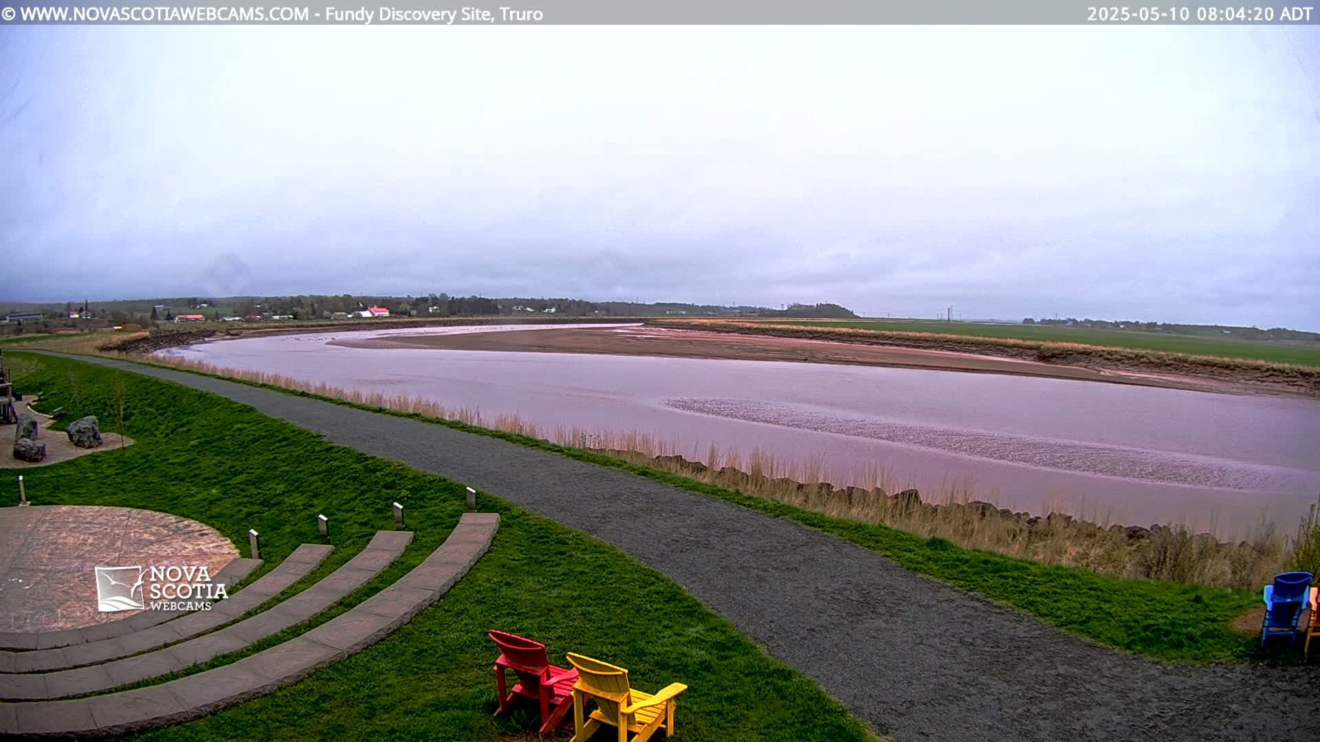 A wide, calm, pinkish-brown river curves gently past a grassy area with a paved path and a few empty chairs under an overcast sky.