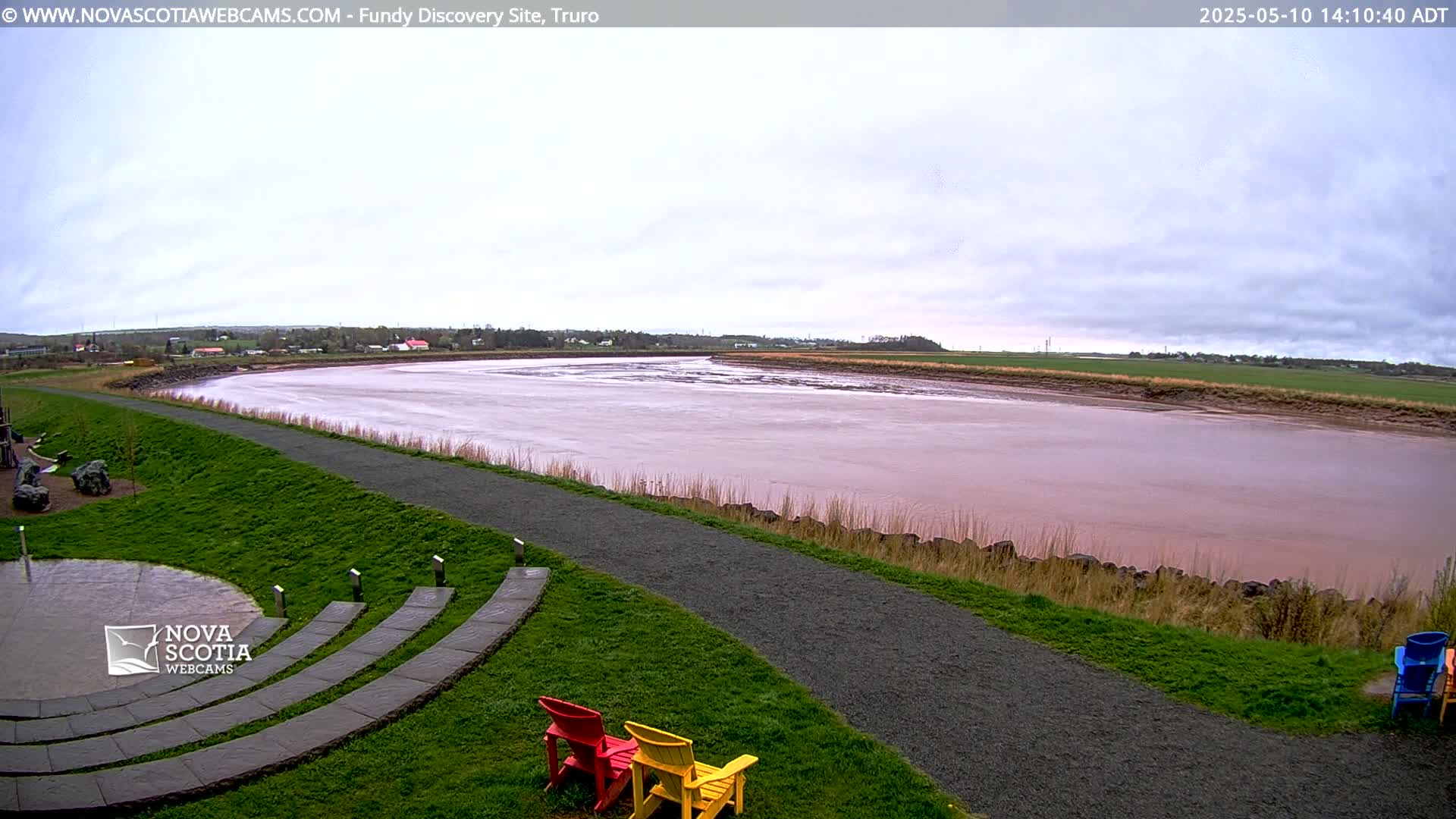 A wide, reddish-brown river flows under a cloudy sky, alongside a paved pathway and grassy area with a few empty chairs.