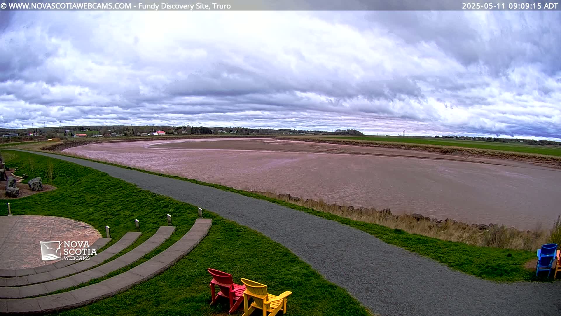 A wide, muddy river stretches out under a mostly cloudy sky, with a paved pathway and grassy area in the foreground, and two colorful chairs near the path.