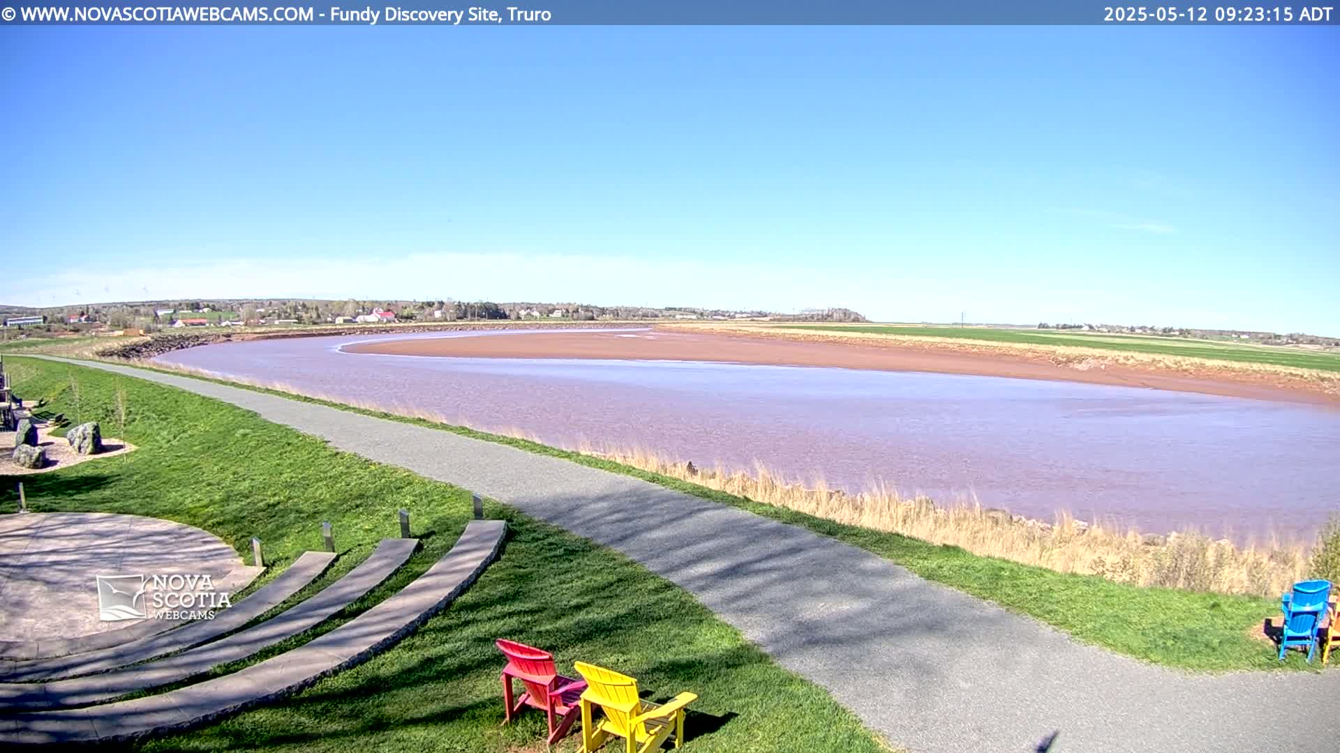 A wide, calm river with reddish-pink water curves past a paved pathway and grassy area with several colorful chairs under a clear blue sky.