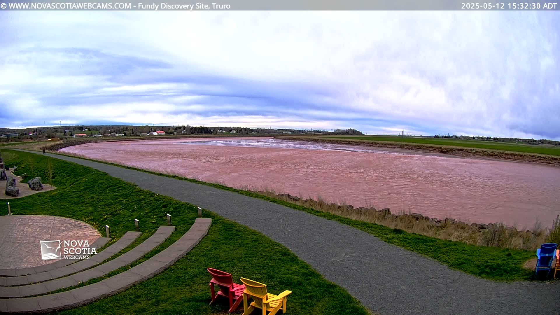 A wide, pink river flows past a paved walkway and grassy area with a few colorful chairs under a partly cloudy sky.
