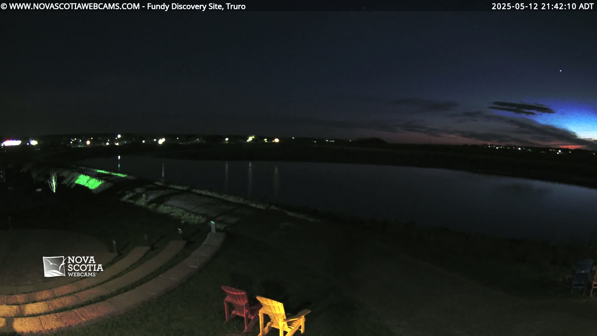 A dark, nighttime scene shows a body of water reflecting lights from a distant shore, with empty Adirondack chairs situated near a stone amphitheater under a partly cloudy sky.