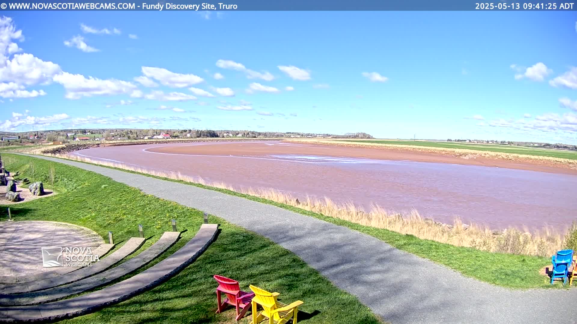 A wide, pinkish-brown river flows under a mostly sunny sky with scattered clouds, beside a paved walkway and grassy area with colorful Adirondack chairs.