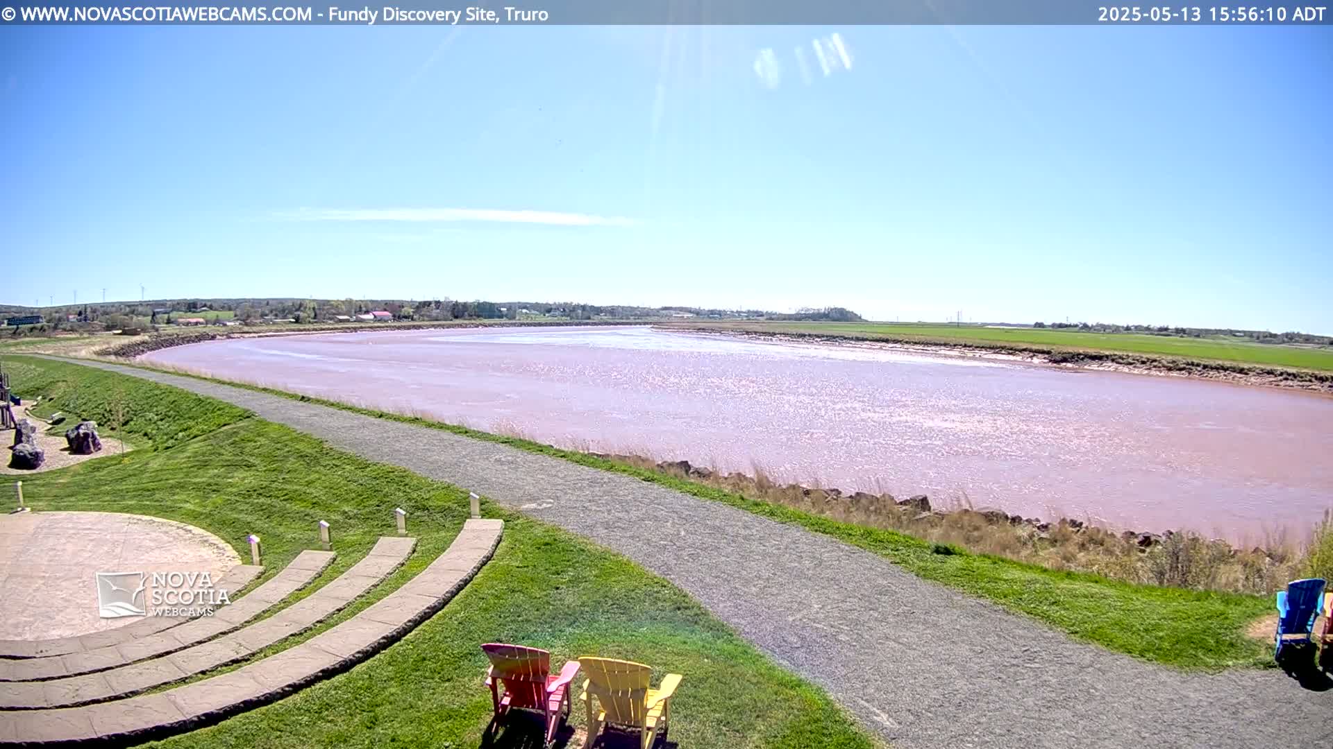A wide, pink-tinged river stretches out under a clear blue sky, alongside a grassy area with a paved pathway and a few Adirondack chairs.