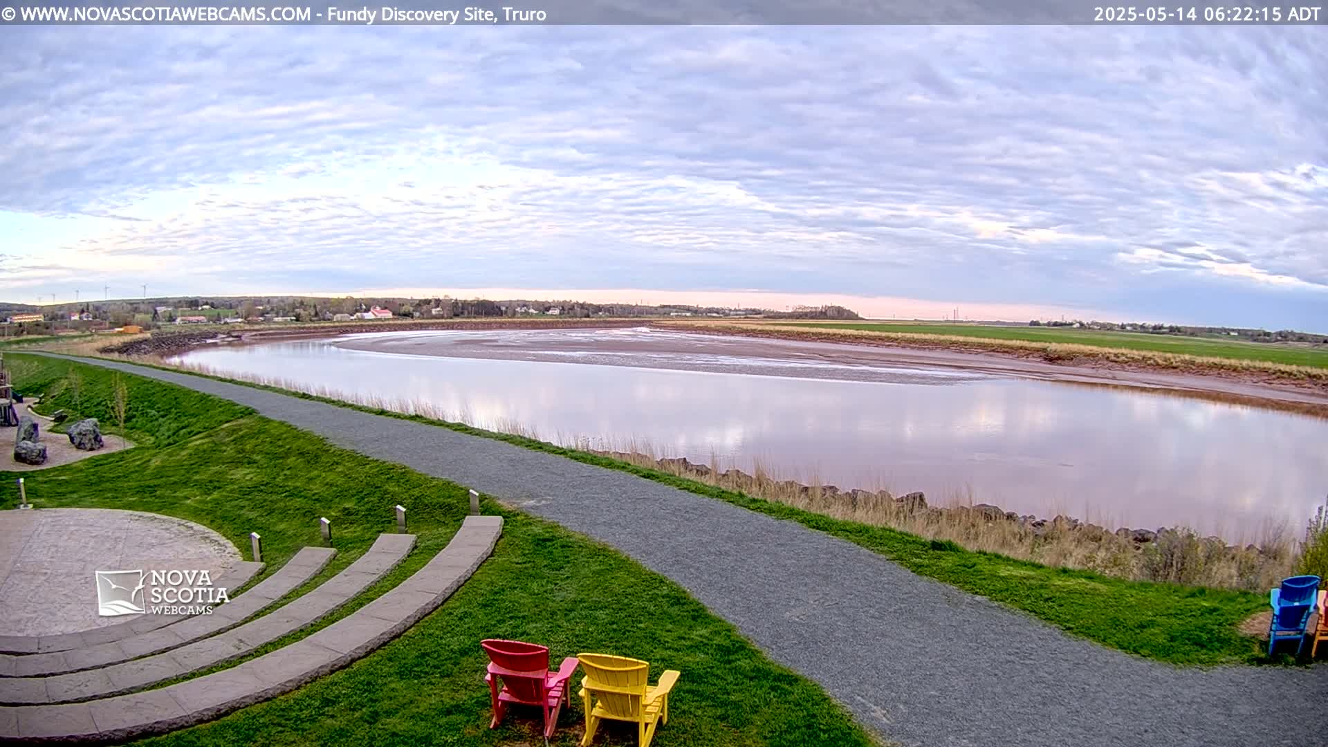 A calm river reflects a cloudy sky, with a grassy area, paved path, and several colorful chairs in the foreground.