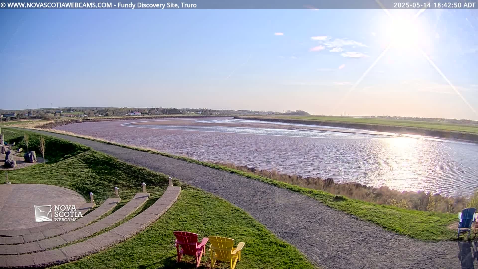 A wide, calm river reflecting sunlight under a mostly clear sky is viewed from a grassy area with two empty chairs and a stone amphitheater.