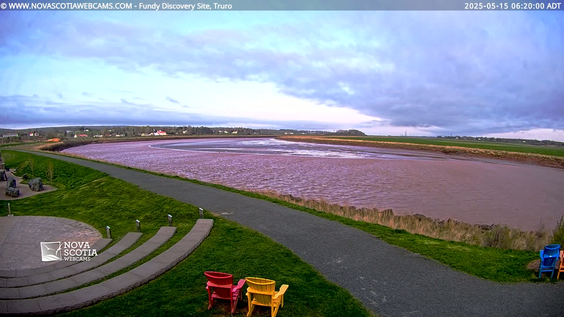 A wide, pinkish-brown river flows under a partly cloudy sky, beside a paved path and grassy area with a few colorful chairs.