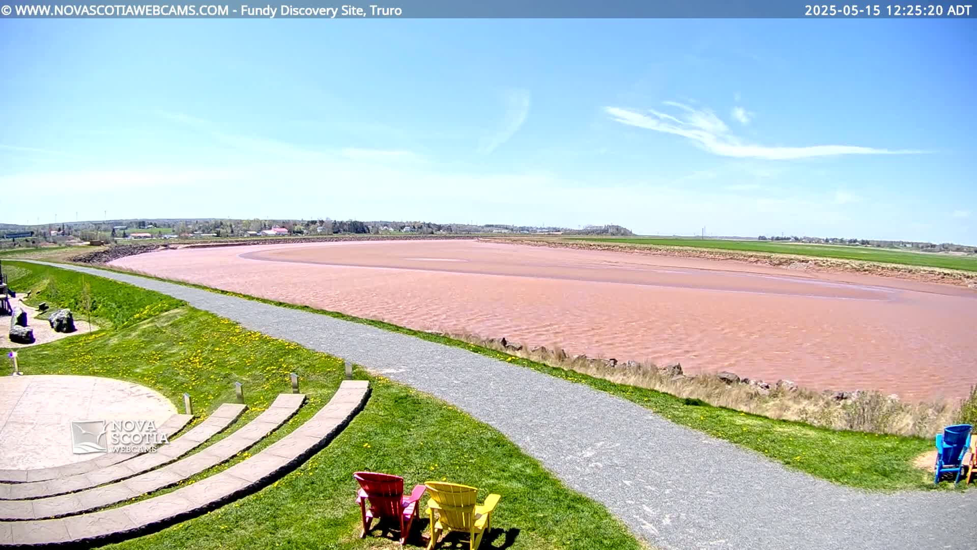 A pinkish-red river flows under a mostly clear blue sky, beside a grassy area with a gravel path and several empty lawn chairs.