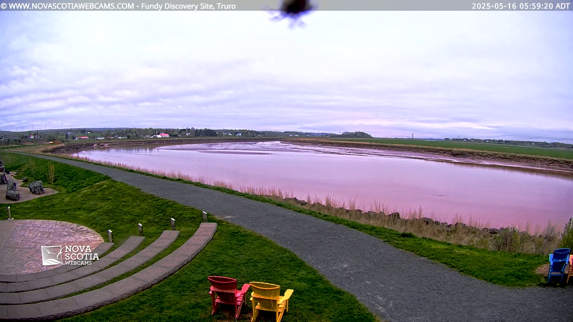 A pinkish-red river flows through a grassy landscape under a cloudy sky, with a paved path and several colorful chairs visible in the foreground.