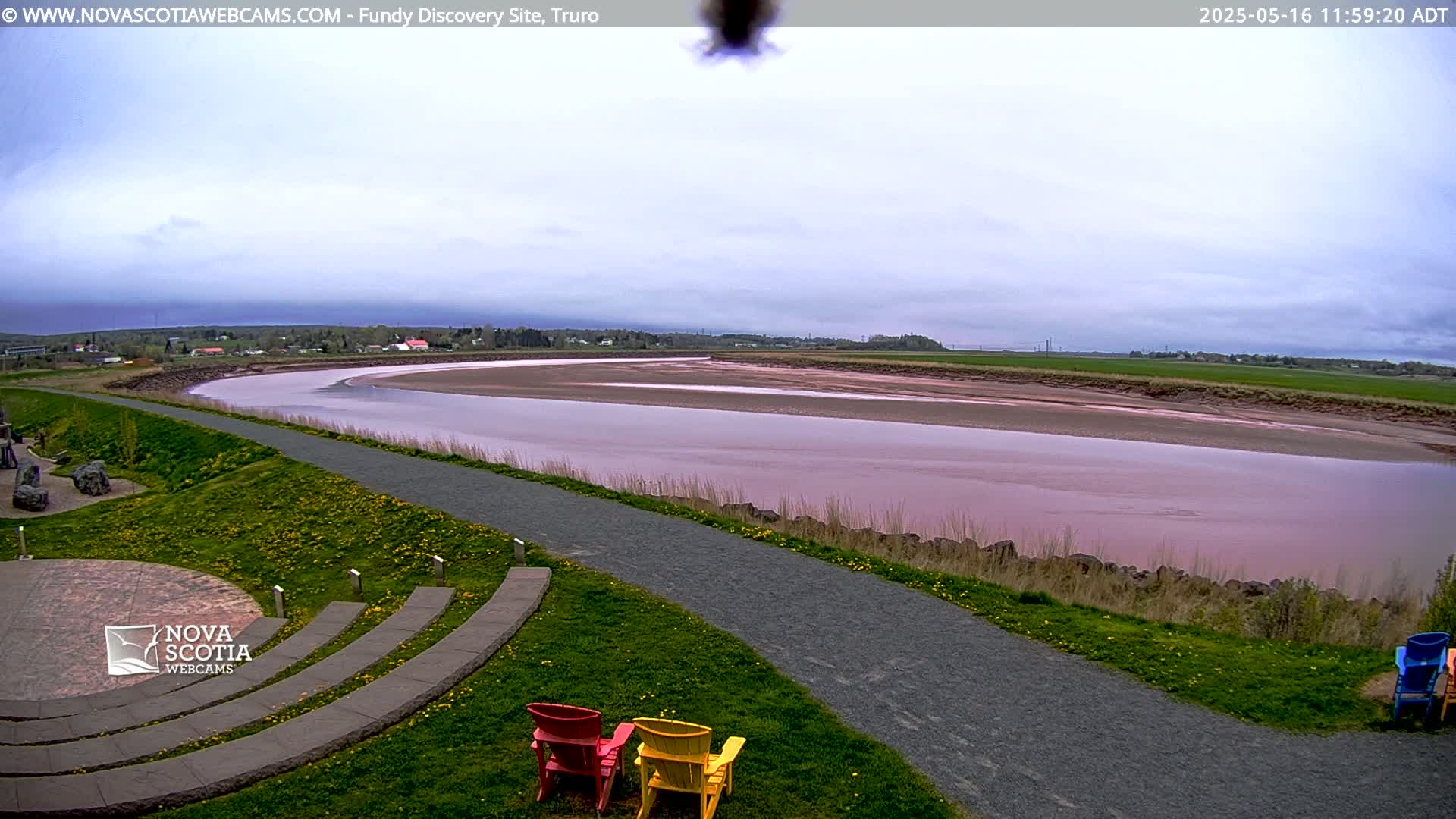 A pinkish-red river winds through a grassy landscape under an overcast sky, with a paved path and several colorful Adirondack chairs in the foreground.